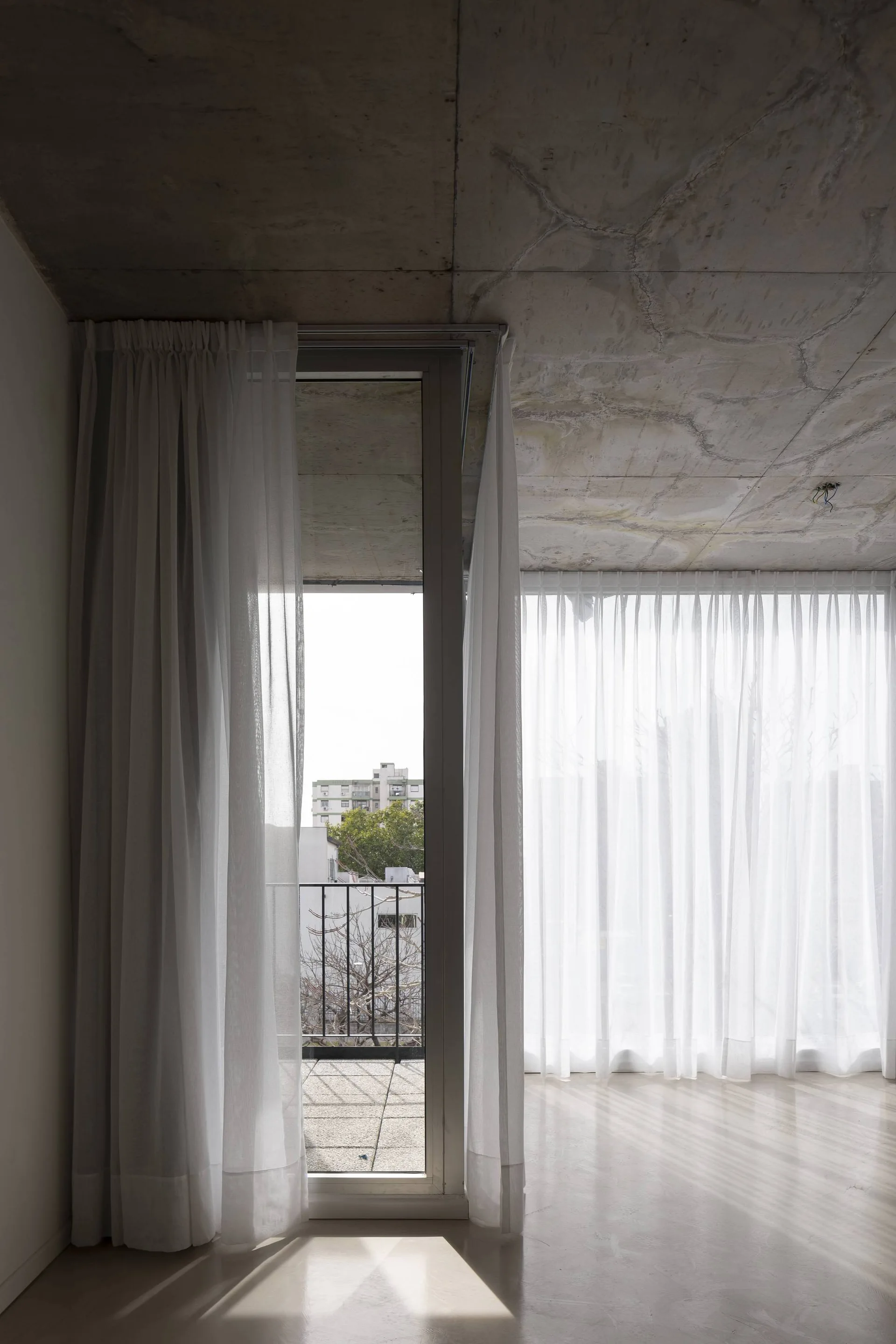 White sheer curtains hang below an exposed raw concrete ceiling in a bright room with a glass door leading to a balcony.