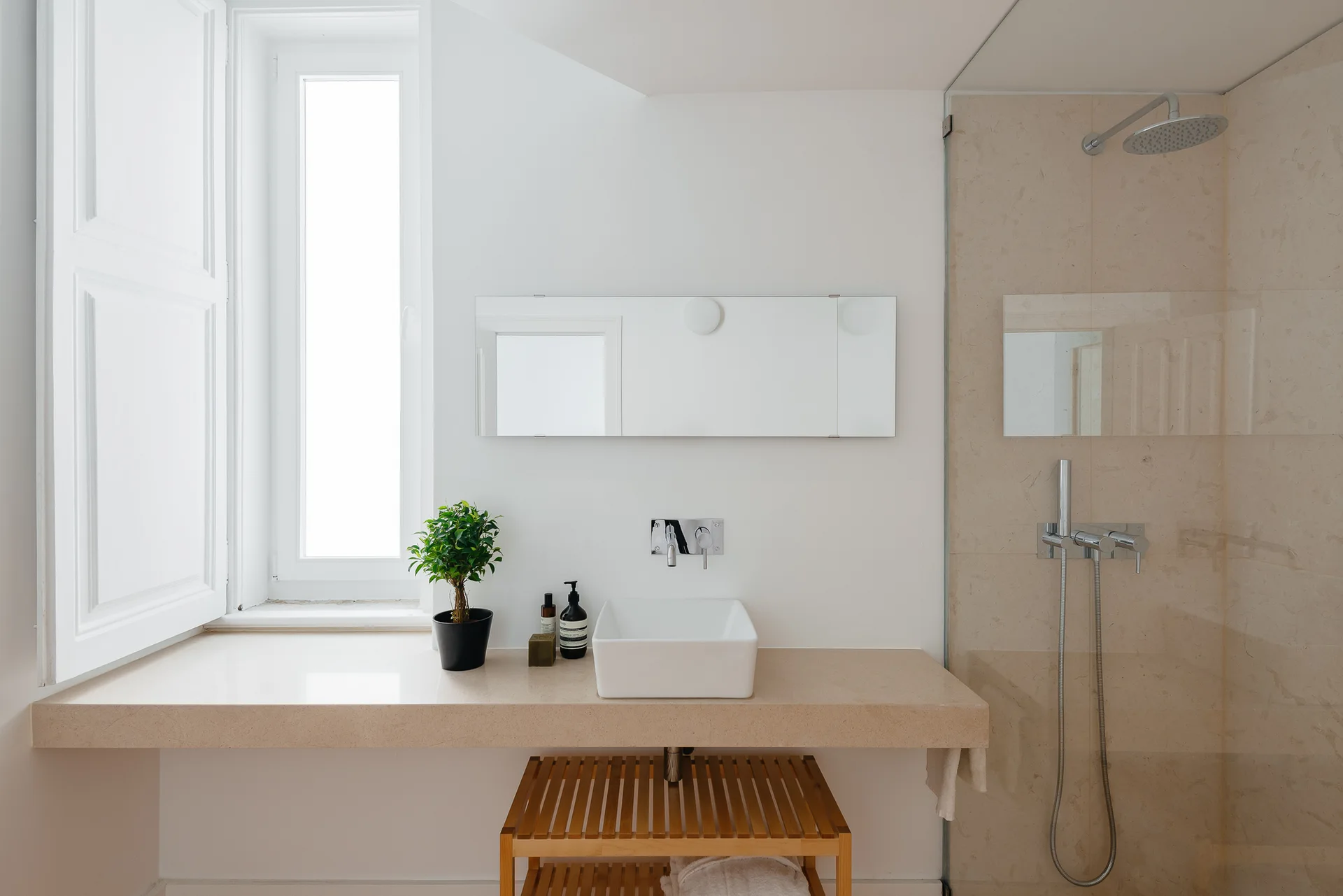 A minimalist bathroom featuring a beige stone vanity with a white vessel sink, a wall-mounted chrome faucet, and a glass-enclosed shower.
