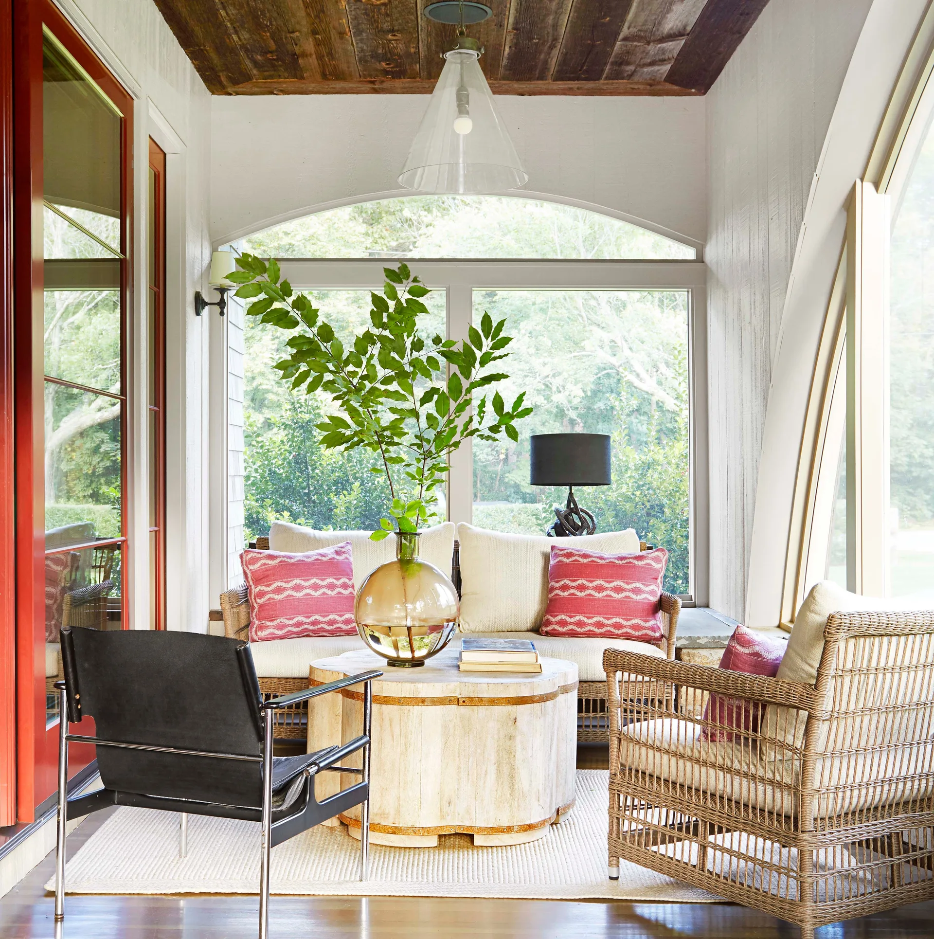 A bright sunroom features a wicker sofa with red patterned pillows, a round wooden coffee table, and a black accent chair beneath a dark plank ceiling.