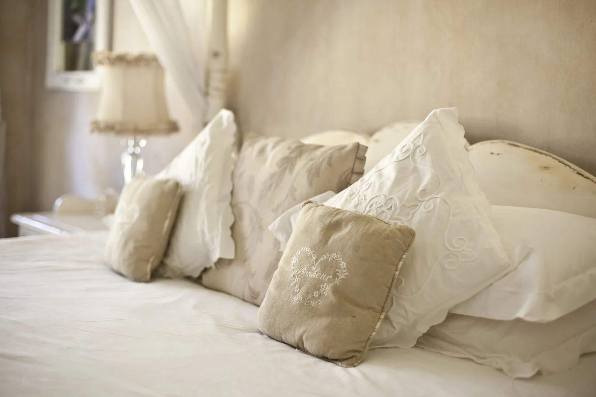 Multiple decorative pillows in cream and beige with delicate embroidery are arranged on a bed in front of a curved, distressed white headboard.
