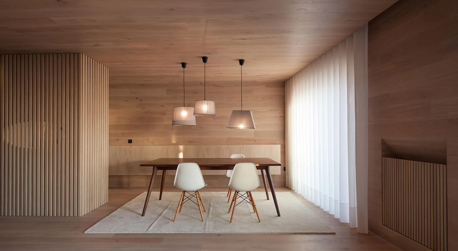 A minimalist dining room featuring light wood wall and ceiling paneling, a dark wood dining table, white chairs, and three pendant lights.