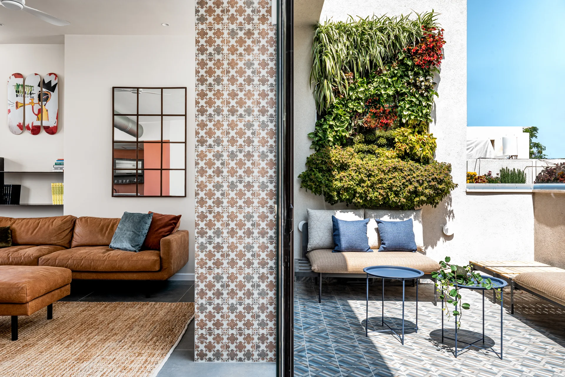 A bright living room with a brown leather sofa transitions to an outdoor patio featuring a lush vertical garden, patterned tiles, and blue metal side tables.