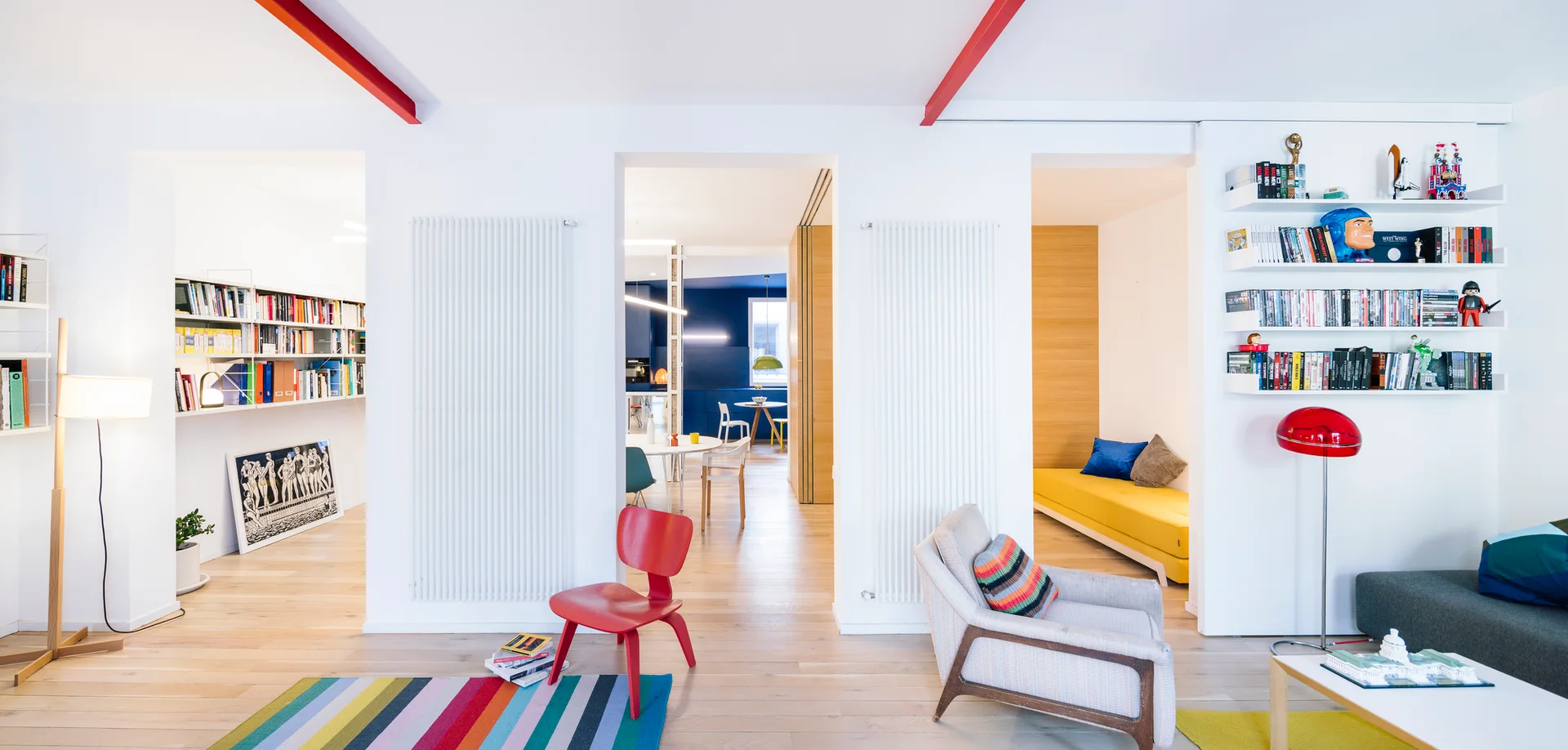 A bright modern living room with light wood floors, red ceiling beams, open doorways leading to other rooms, and a colorful striped rug.