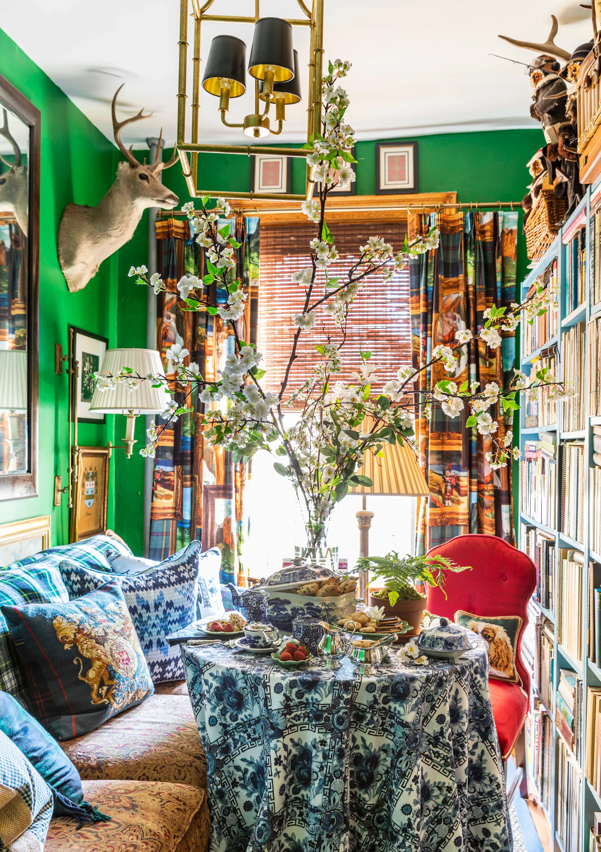 A small dining table with a blue floral tablecloth sits in a green room next to a tall bookshelf filled with books.