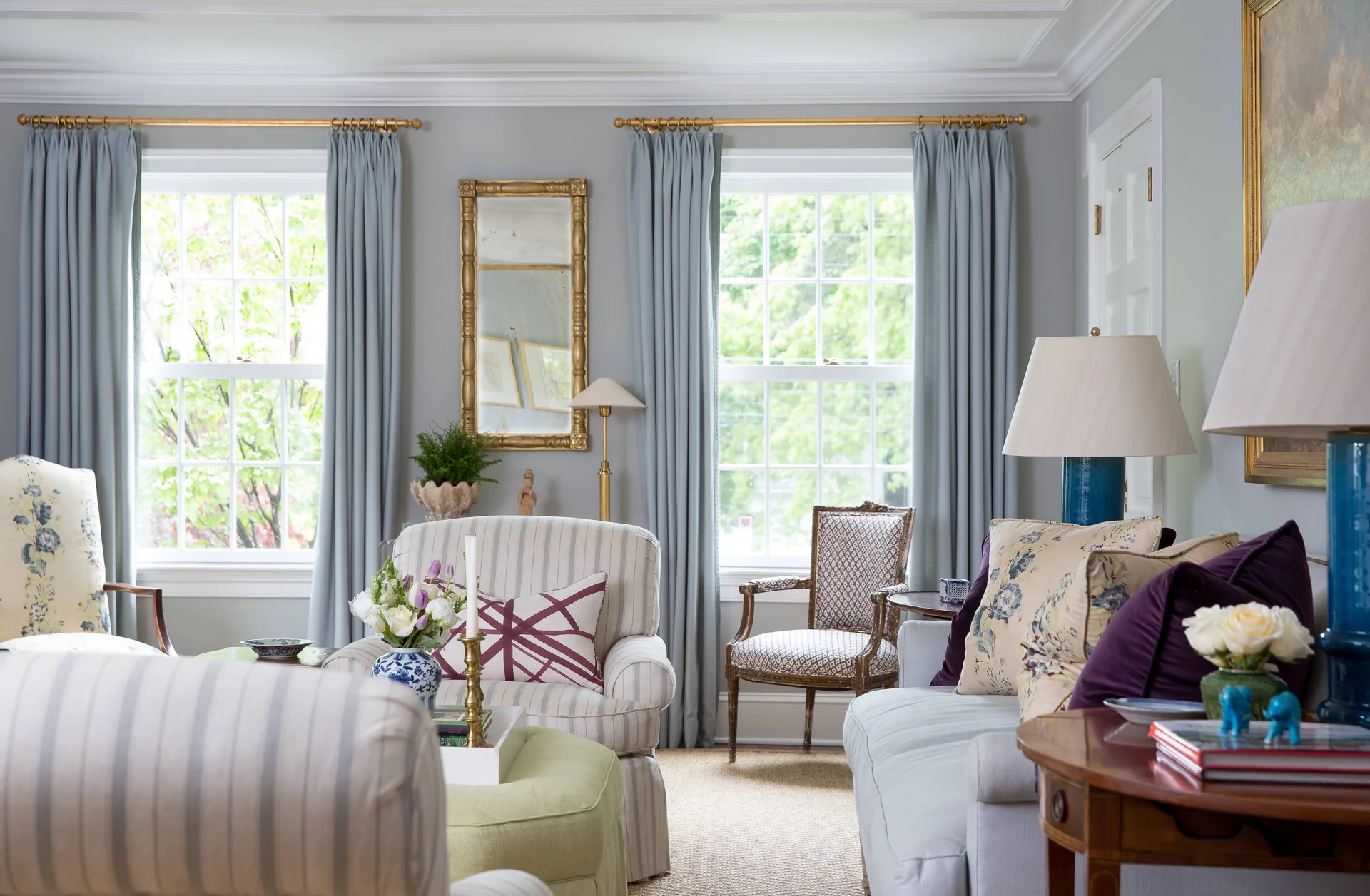 A traditional living room featuring striped armchairs, a green ottoman, blue curtains, and a gold-framed mirror mounted between two windows.