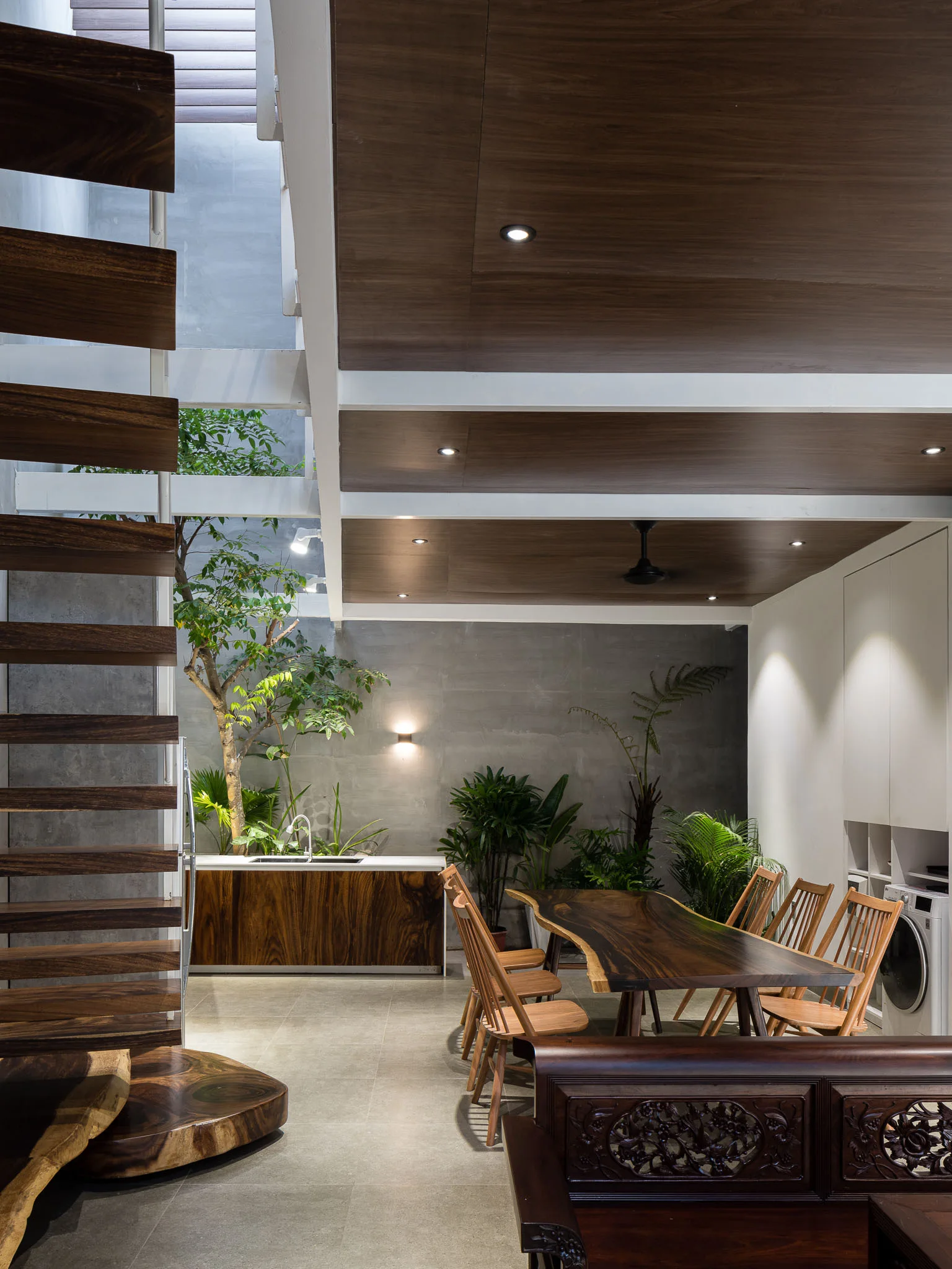 An open-concept dining area featuring a live-edge wooden table, an open-riser staircase, an indoor plant garden, and concrete walls.