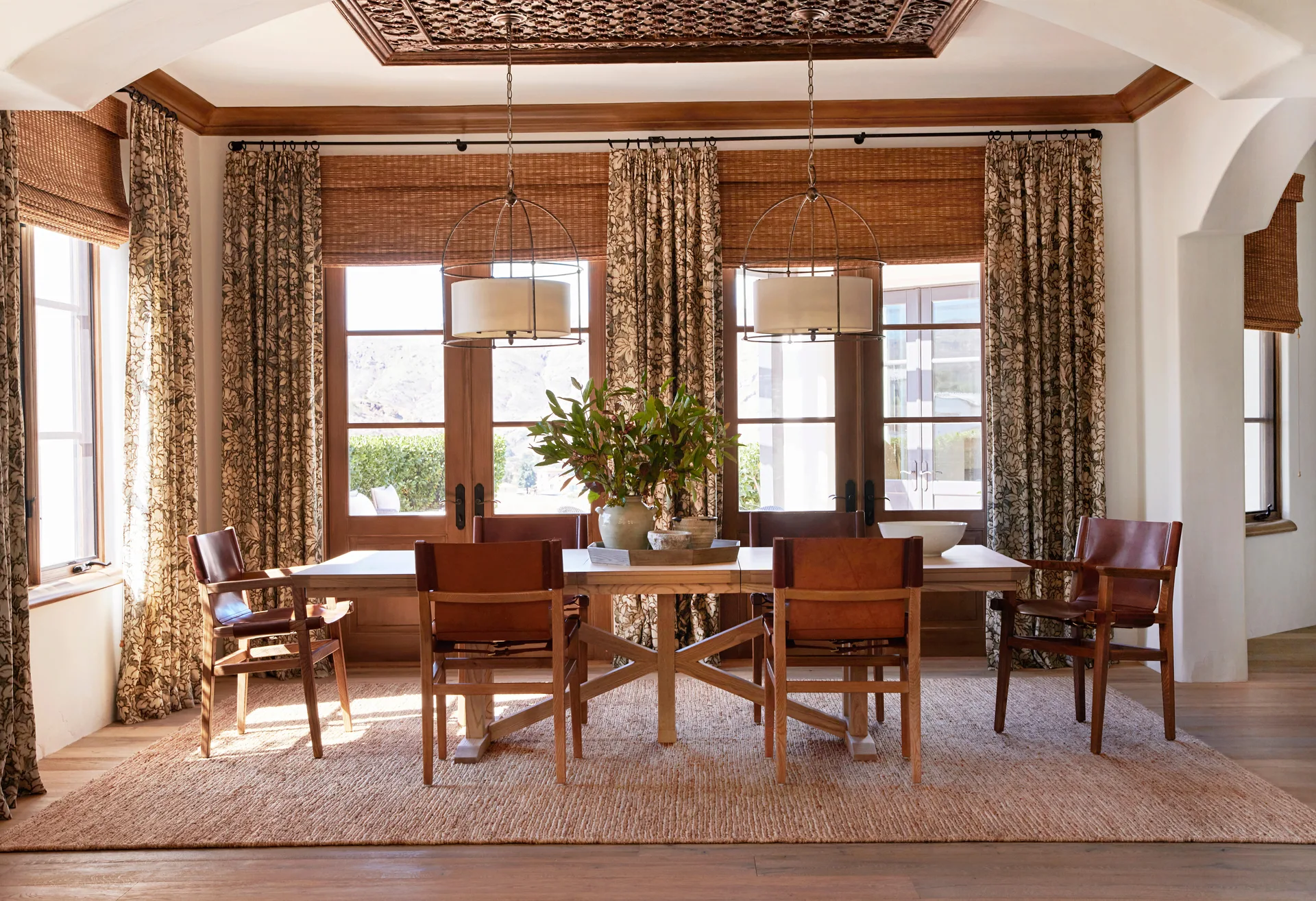 A sunlit Mediterranean style dining room featuring a long wooden table, leather and wood chairs, botanical print curtains, and woven roman shades.
