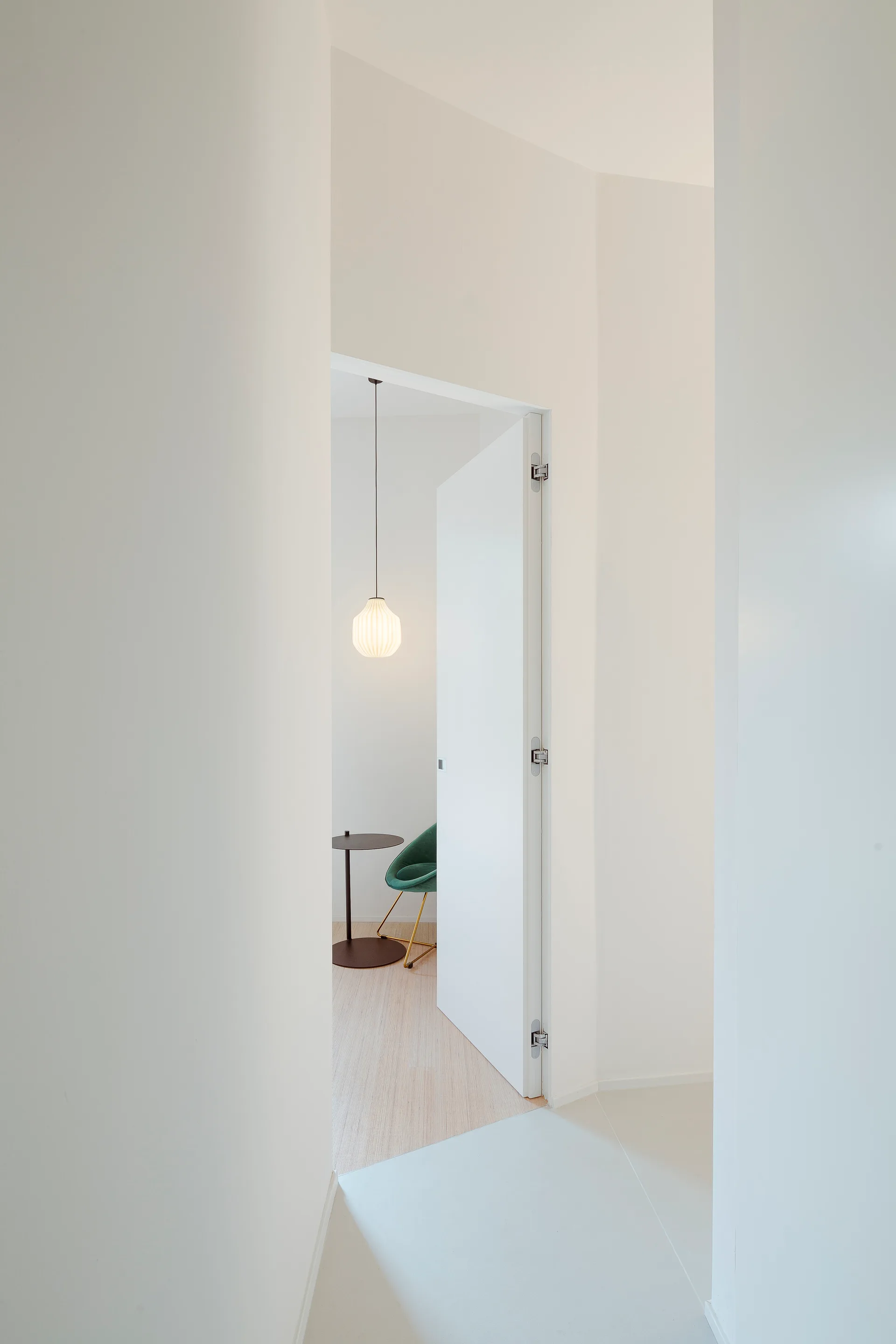 A view through an open white doorway shows a minimalist room with a green velvet chair, a small pedestal table, and a glowing ribbed pendant light.