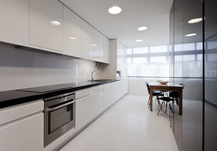 A minimalist galley kitchen featuring high-gloss white handleless cabinets, a black countertop, and a wood dining table with chairs by a window.