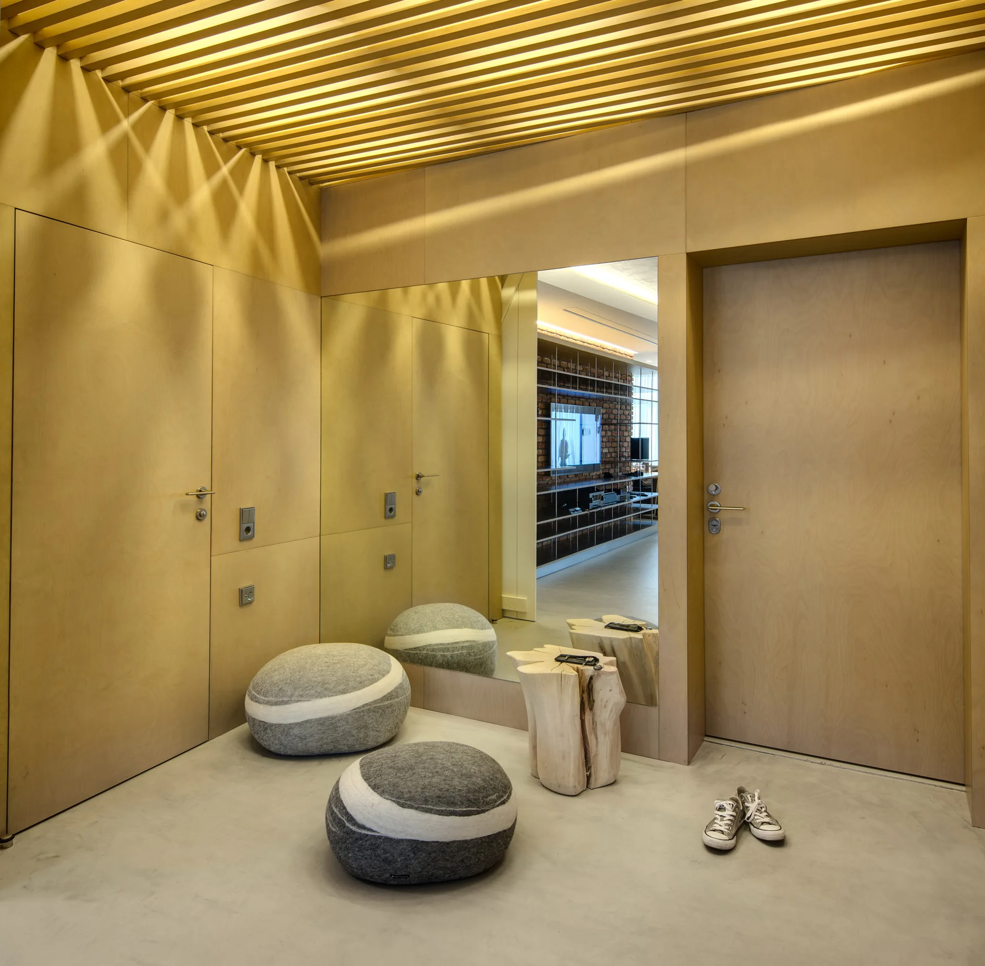 A minimalist entryway features light wood wall paneling, a slatted ceiling, a large wall mirror, and grey pebble-shaped cushions on a concrete floor.