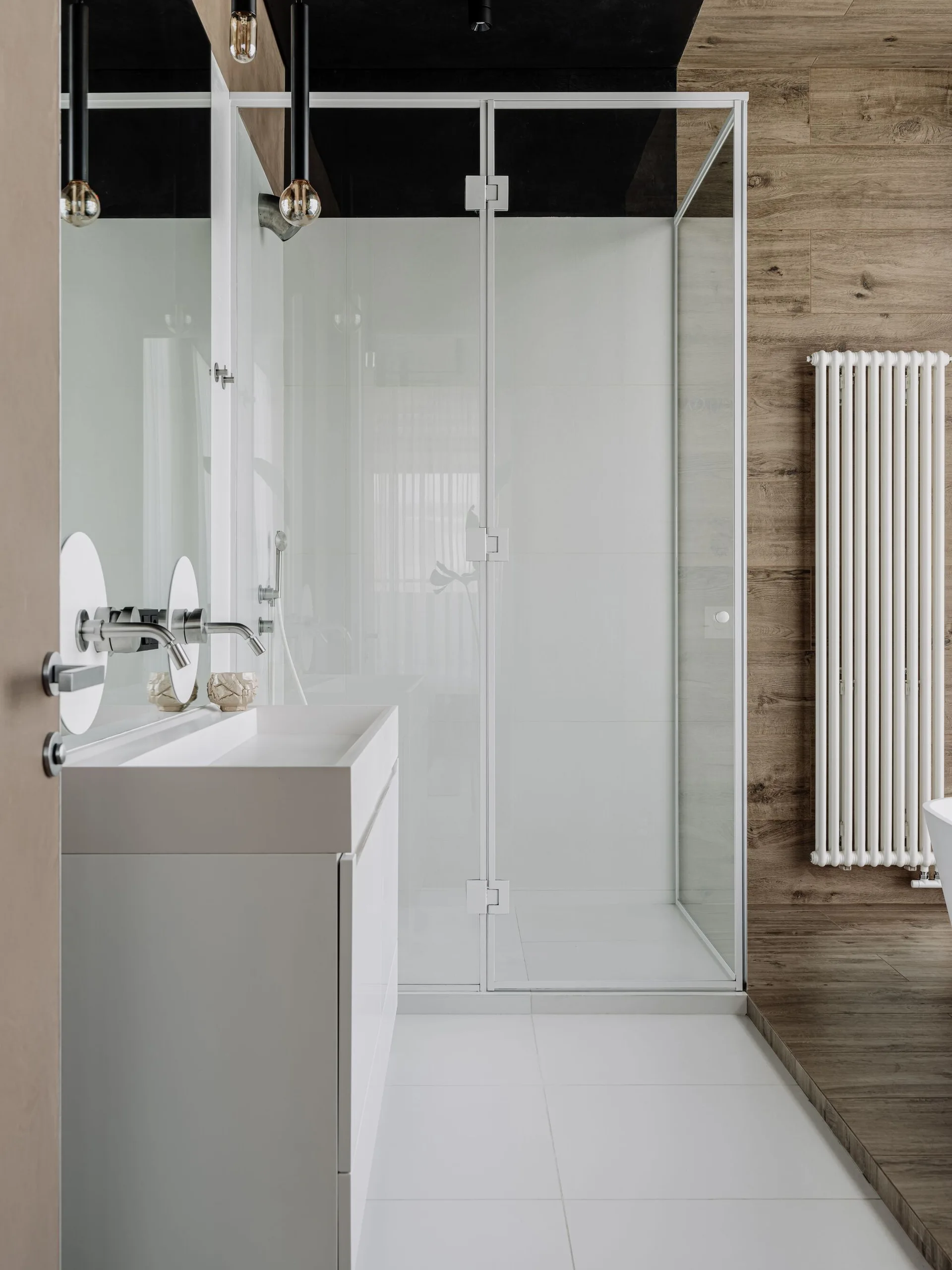 A modern bathroom showing a white vanity with wall-mounted faucets, a glass shower enclosure, and a white vertical radiator against a wood-plank wall.