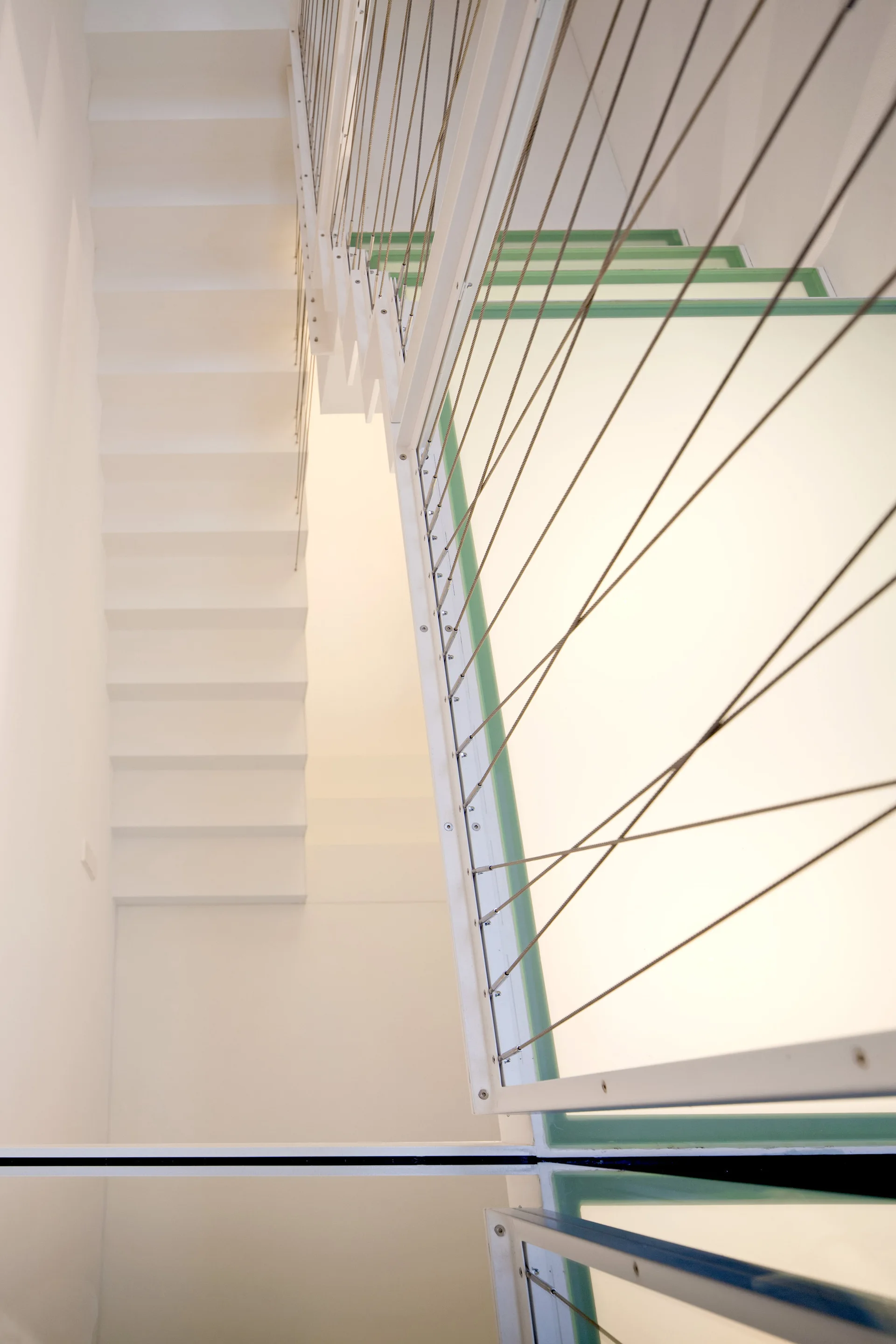 A high-angle view of a modern white staircase featuring metal railings with tension cables and transparent glass-edged treads.
