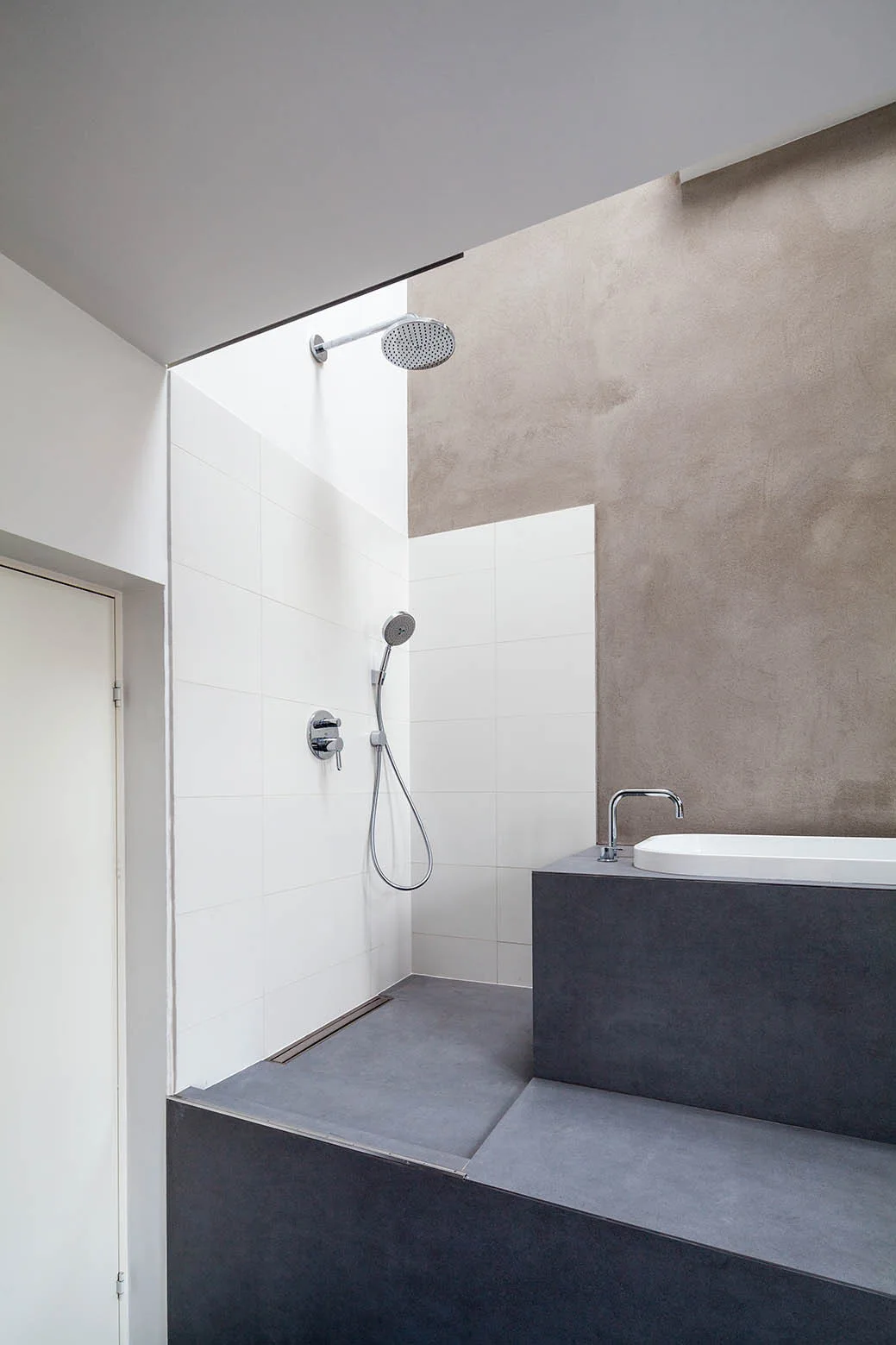A modern bathroom featuring a raised dark grey bathtub platform next to a shower area with white square tiles and a textured brown plaster wall.