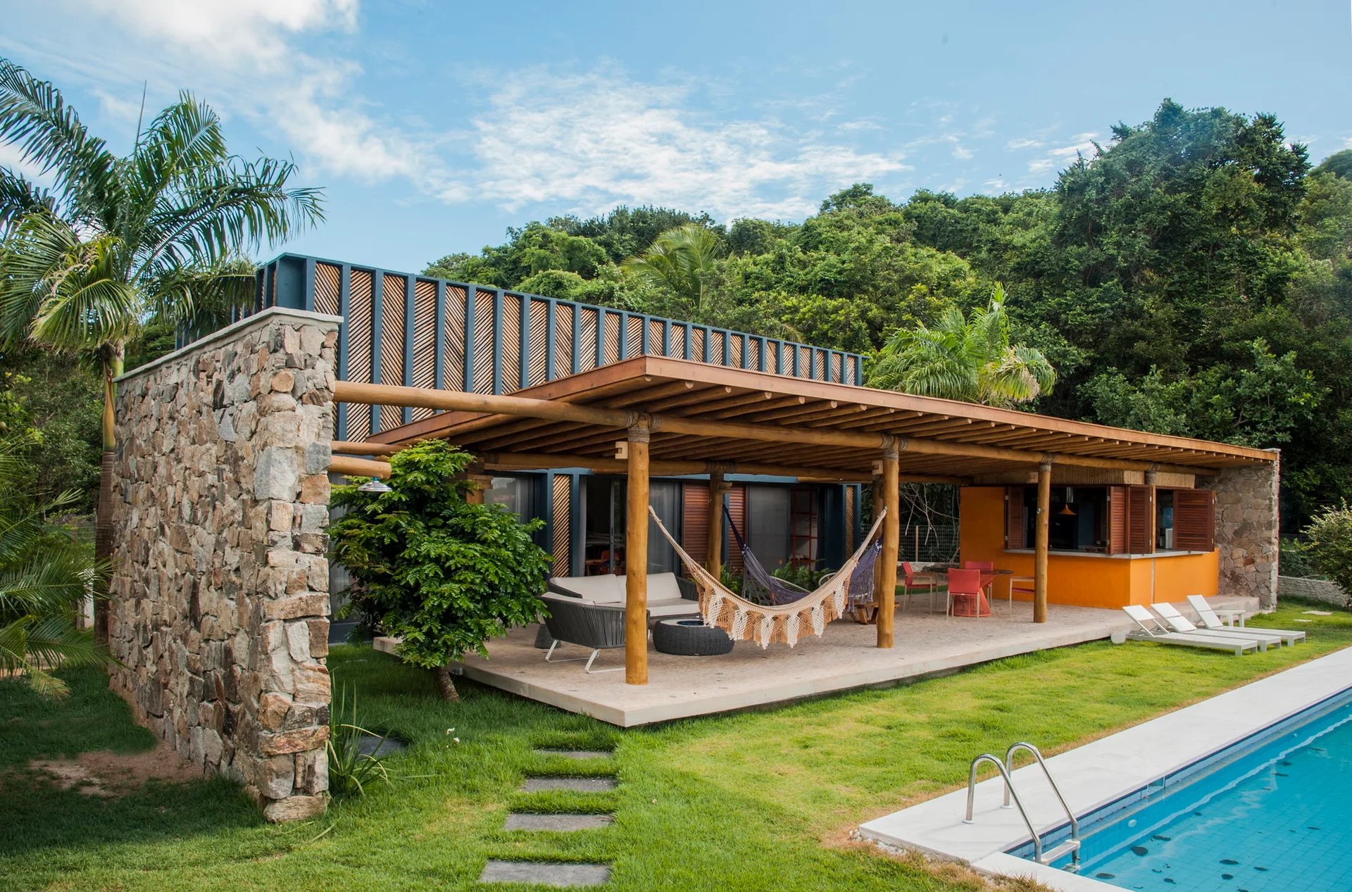 A covered tropical patio features a wooden pergola ceiling, a hanging fringed hammock, and a stone feature wall next to a swimming pool.