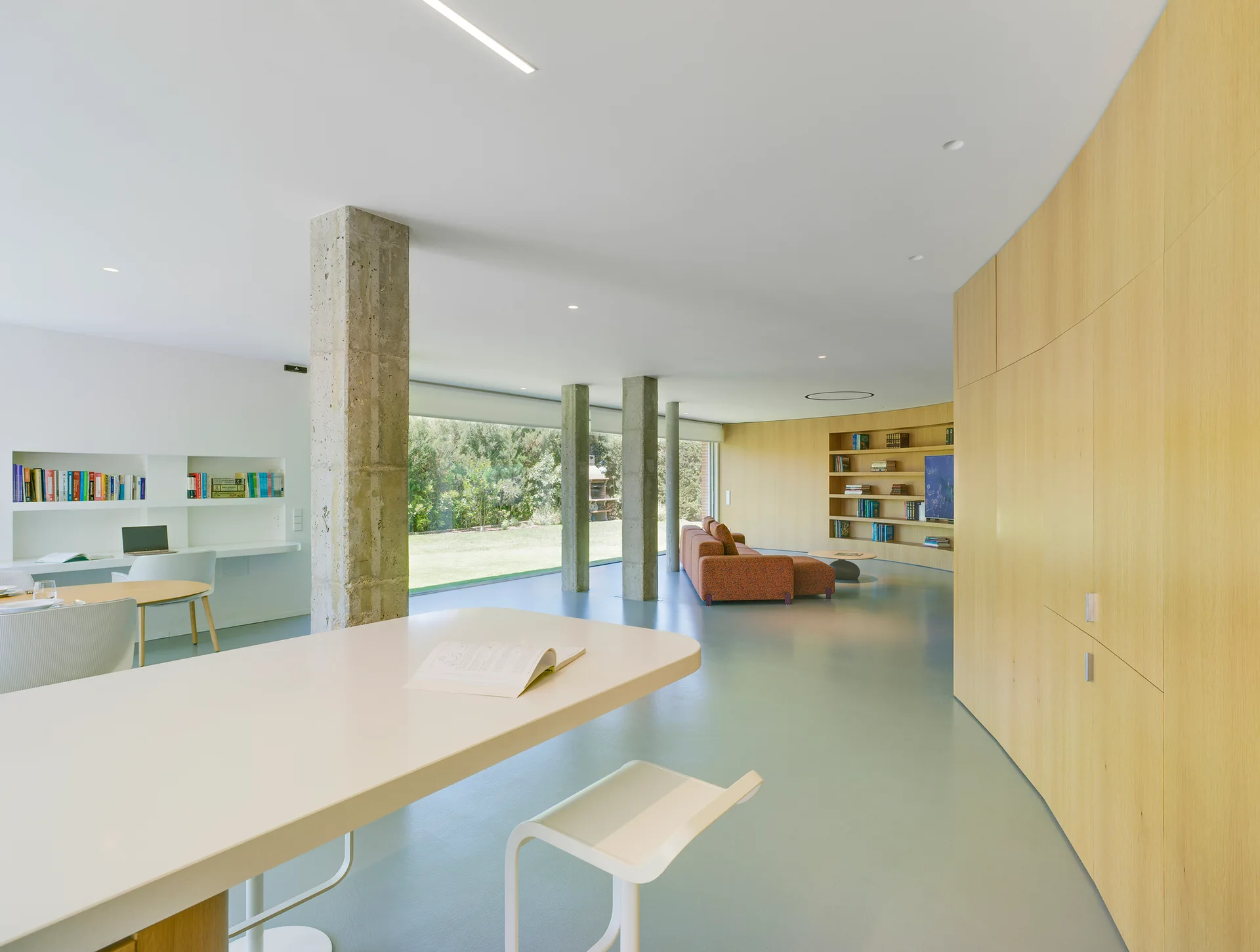A minimalist open-plan interior features a curved light wood storage wall, exposed concrete columns, and a work area next to large garden windows.