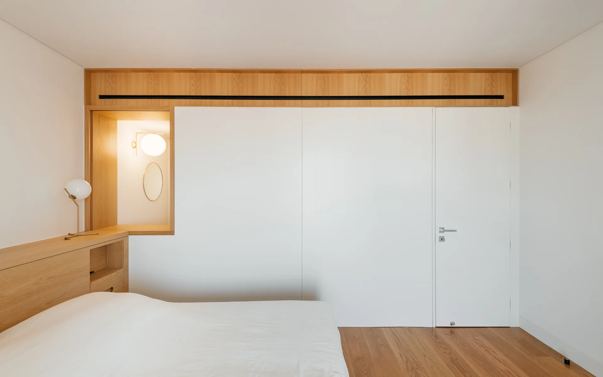 A minimalist bedroom features custom white closet doors framed in light oak wood, a built-in headboard ledge, and a recessed lighted niche.