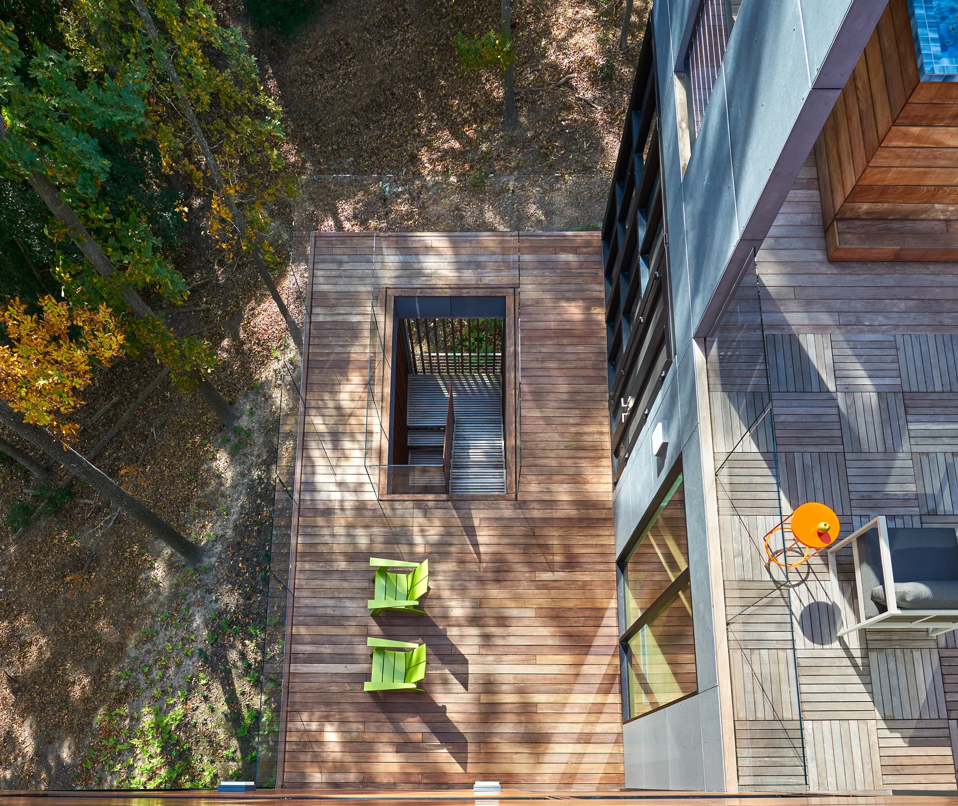 An aerial view of a multi-level wood deck with a recessed central staircase, glass railings, lime green chairs, and surrounding forest trees.