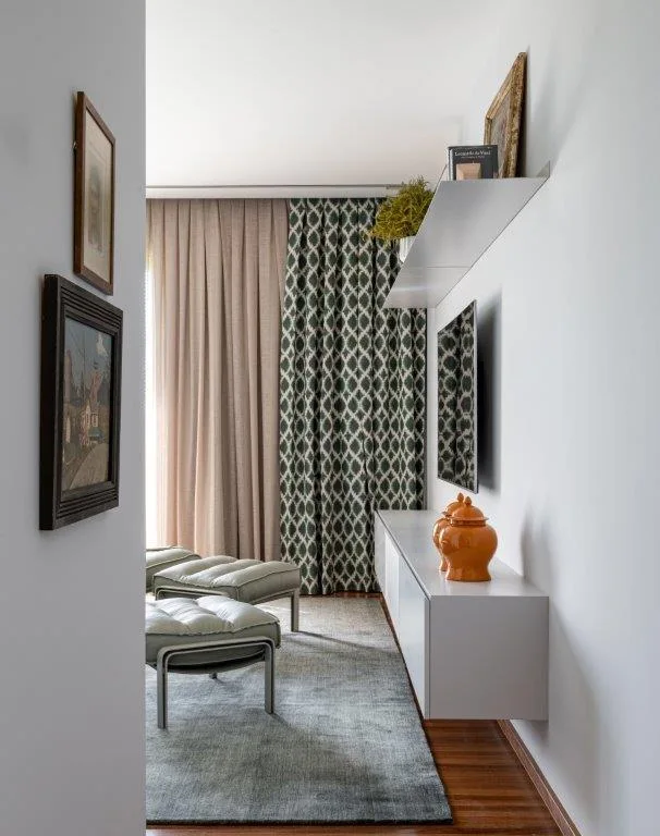 A modern living area features a white floating media console, a pair of green upholstered ottomans, and patterned drapes next to solid curtains.