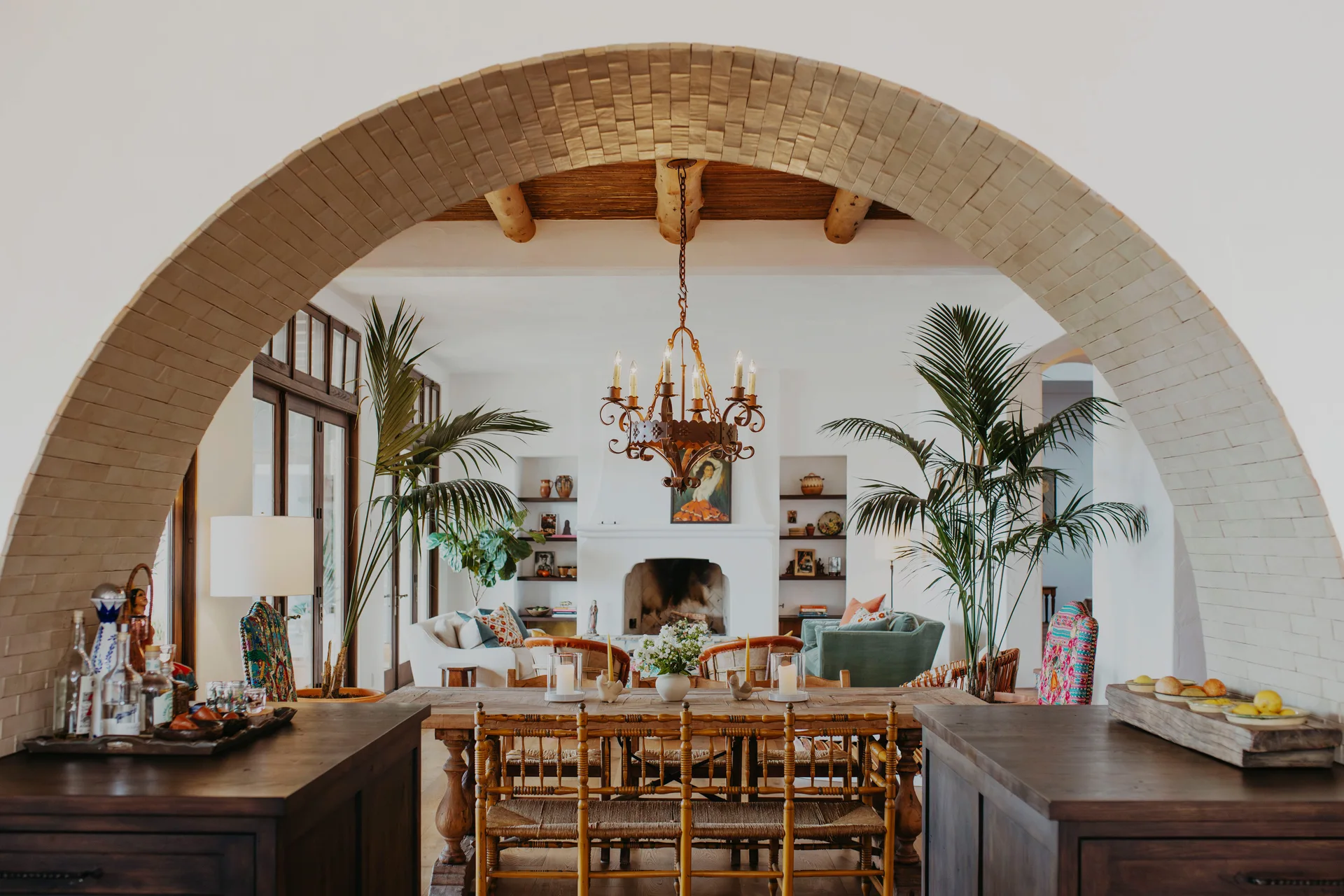 A rustic wooden dining table and spindle chairs sit beneath a wrought iron chandelier, viewed through a large brick-lined archway.