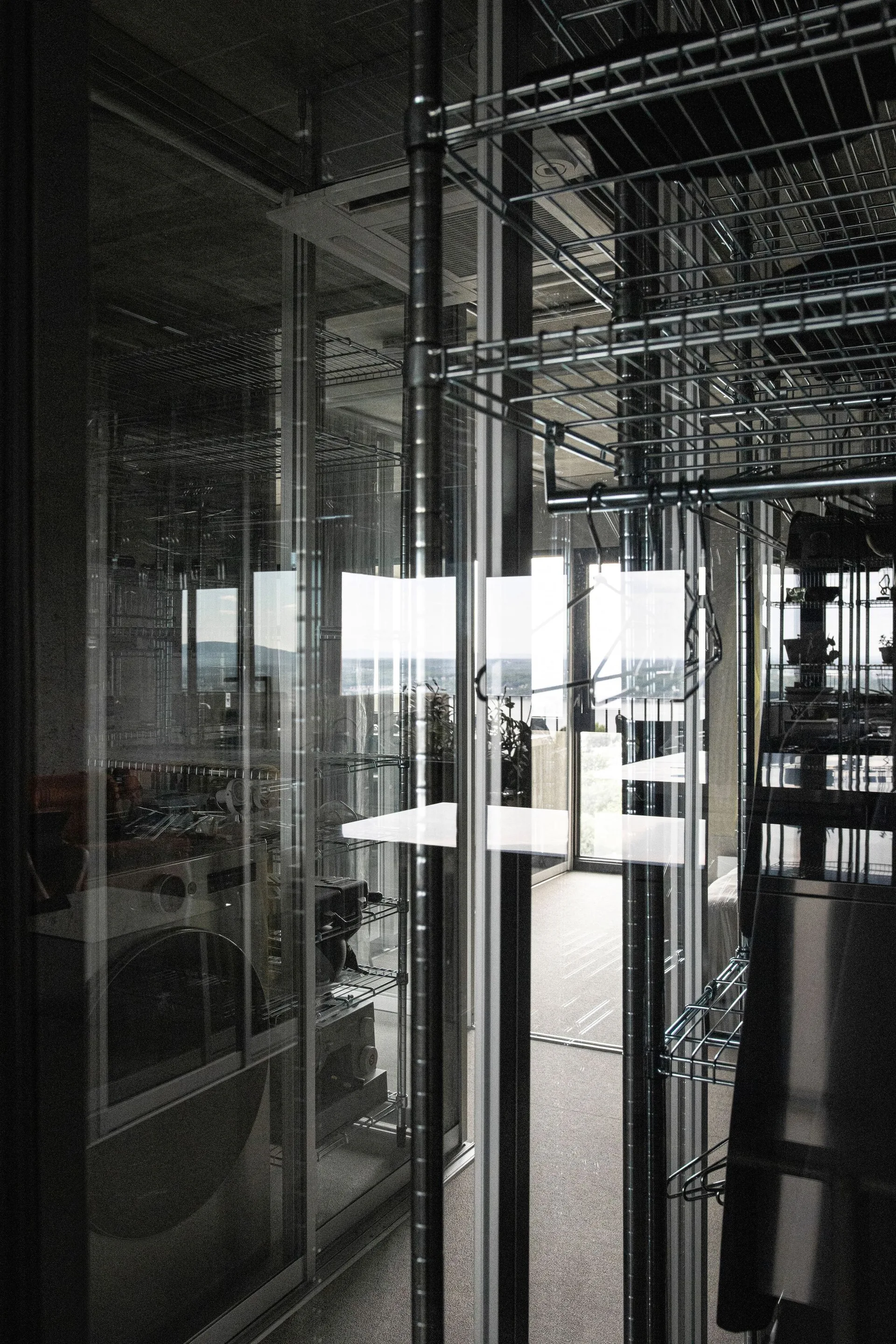 An industrial utility room features metal wire shelving units behind large glass partitions, with concrete ceilings and a view toward an outdoor glass door.