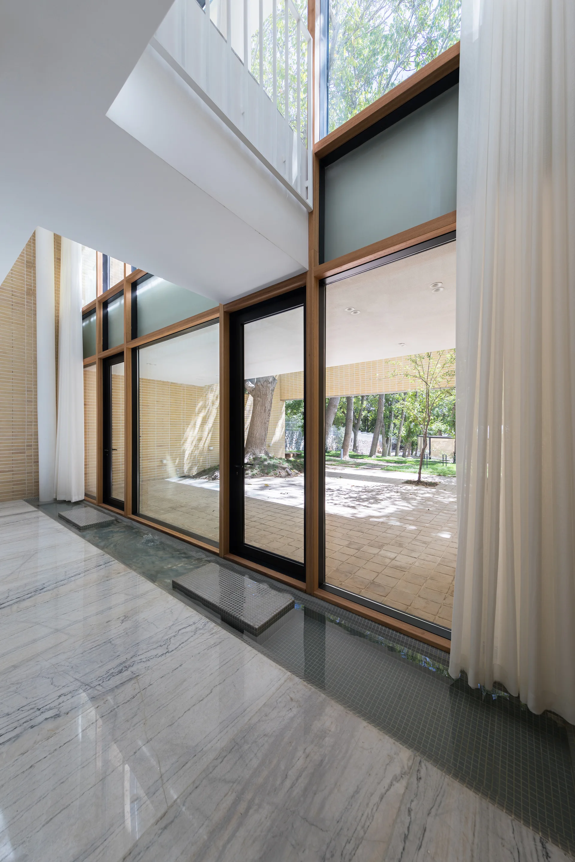 A modern interior entryway features veined white marble floors, an indoor tiled water feature running along wood-framed glass doors, and sheer white curtains.