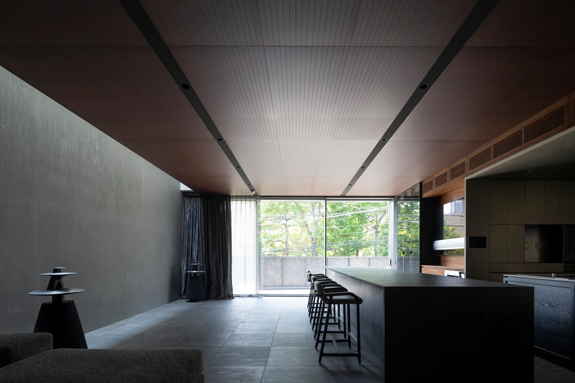 A dark minimalist kitchen features a long black stone island with bar stools, a wood plank ceiling, and floor-to-ceiling glass doors opening to trees.