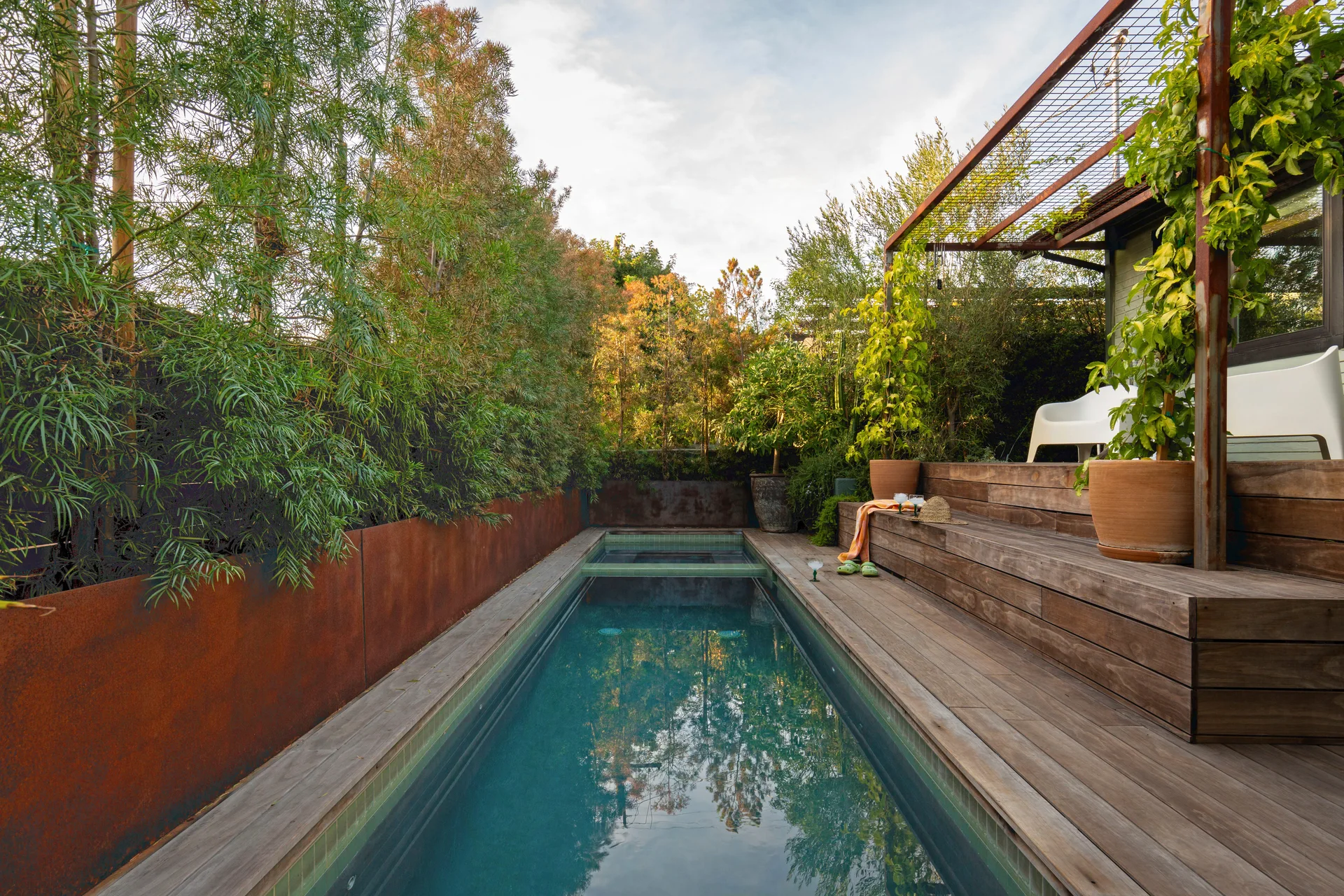 A long rectangular swimming pool sits between multi-level wooden decking and a rusted metal retaining wall topped with lush green trees.