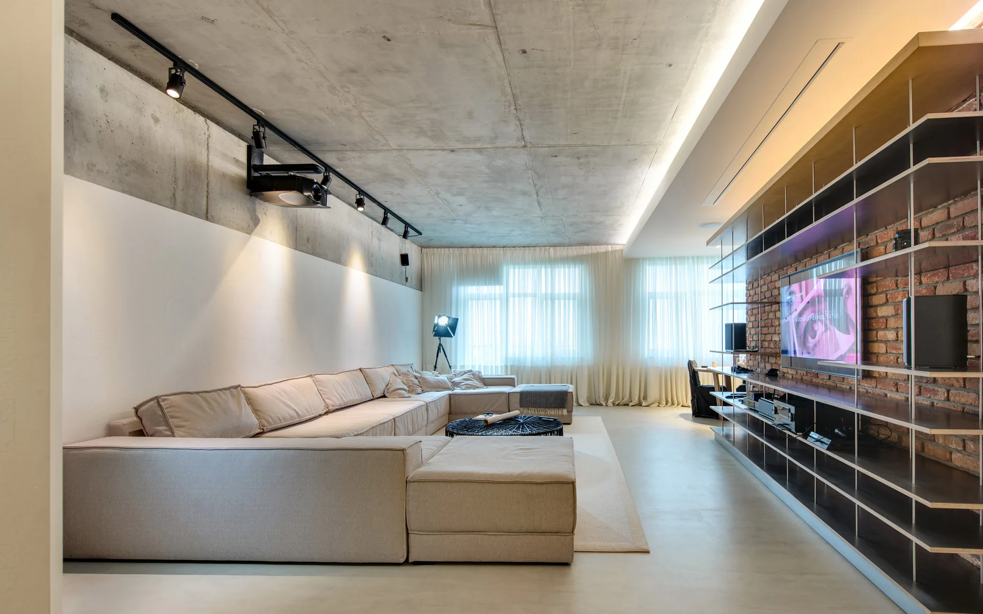 A spacious industrial living room features a large beige sectional sofa, a raw concrete ceiling, and a television mounted on an exposed brick wall.