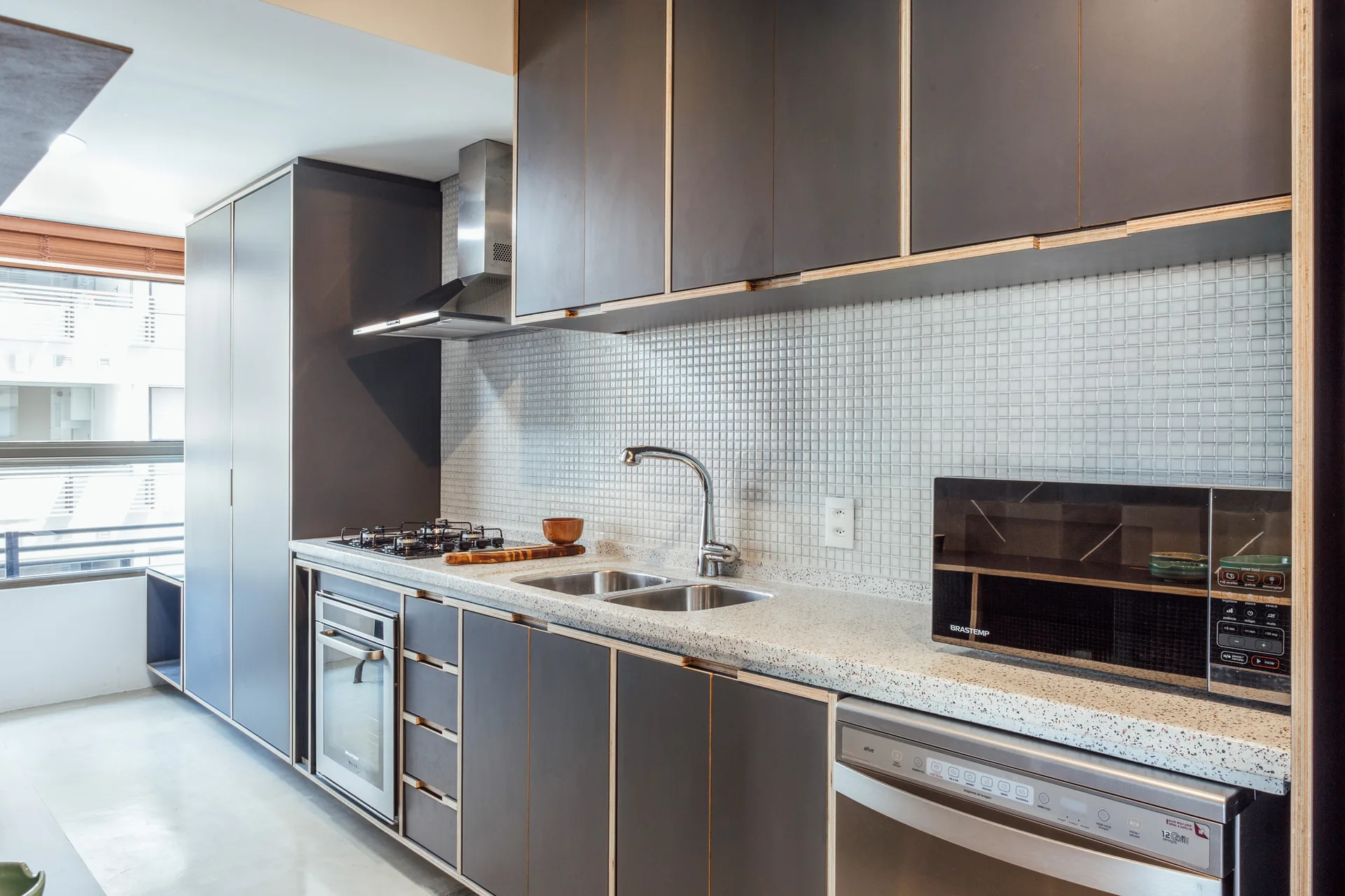 Modern kitchen with dark grey cabinets showing exposed plywood edges, a speckled countertop, and a white mosaic tile backsplash.