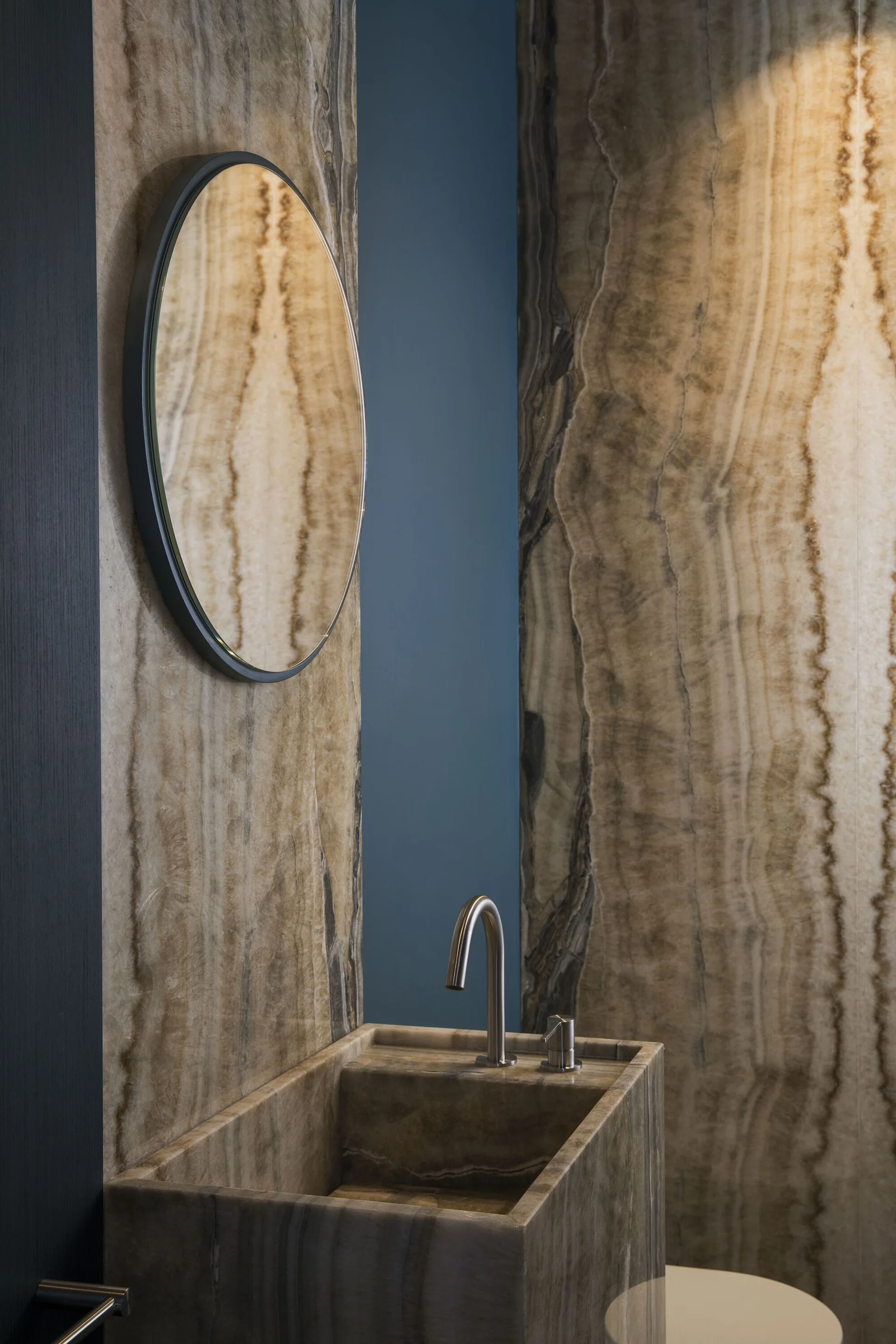 A modern bathroom features a rectangular stone sink basin set against matching veined stone wall panels and a round black-framed mirror.
