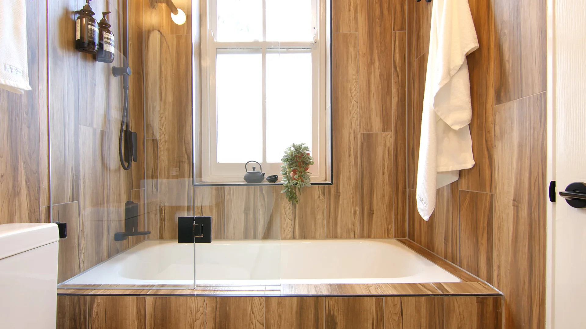 A white inset bathtub is surrounded by vertical wood-look ceramic tiles, featuring a clear glass shower screen and a central sash window.