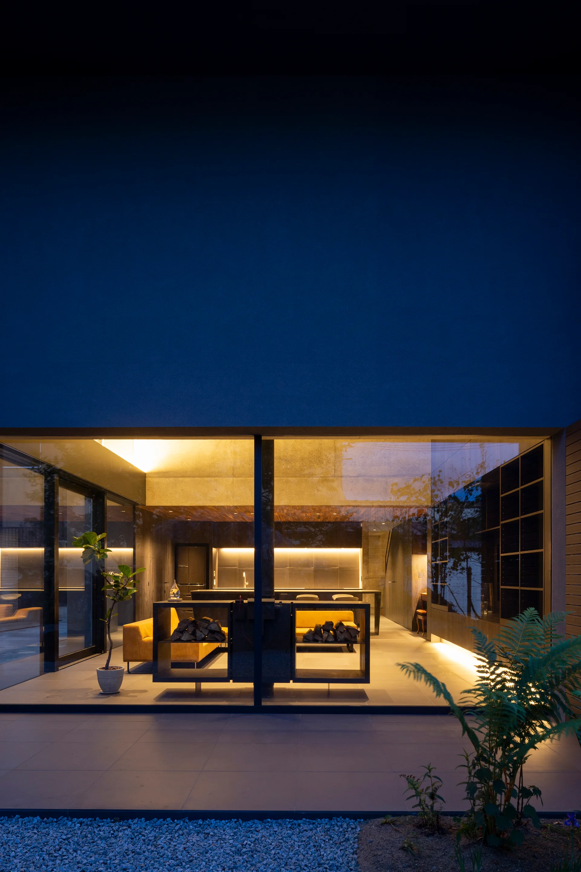 A view through a large glass wall into a modern living room at night, featuring a yellow sofa and warm indirect lighting.