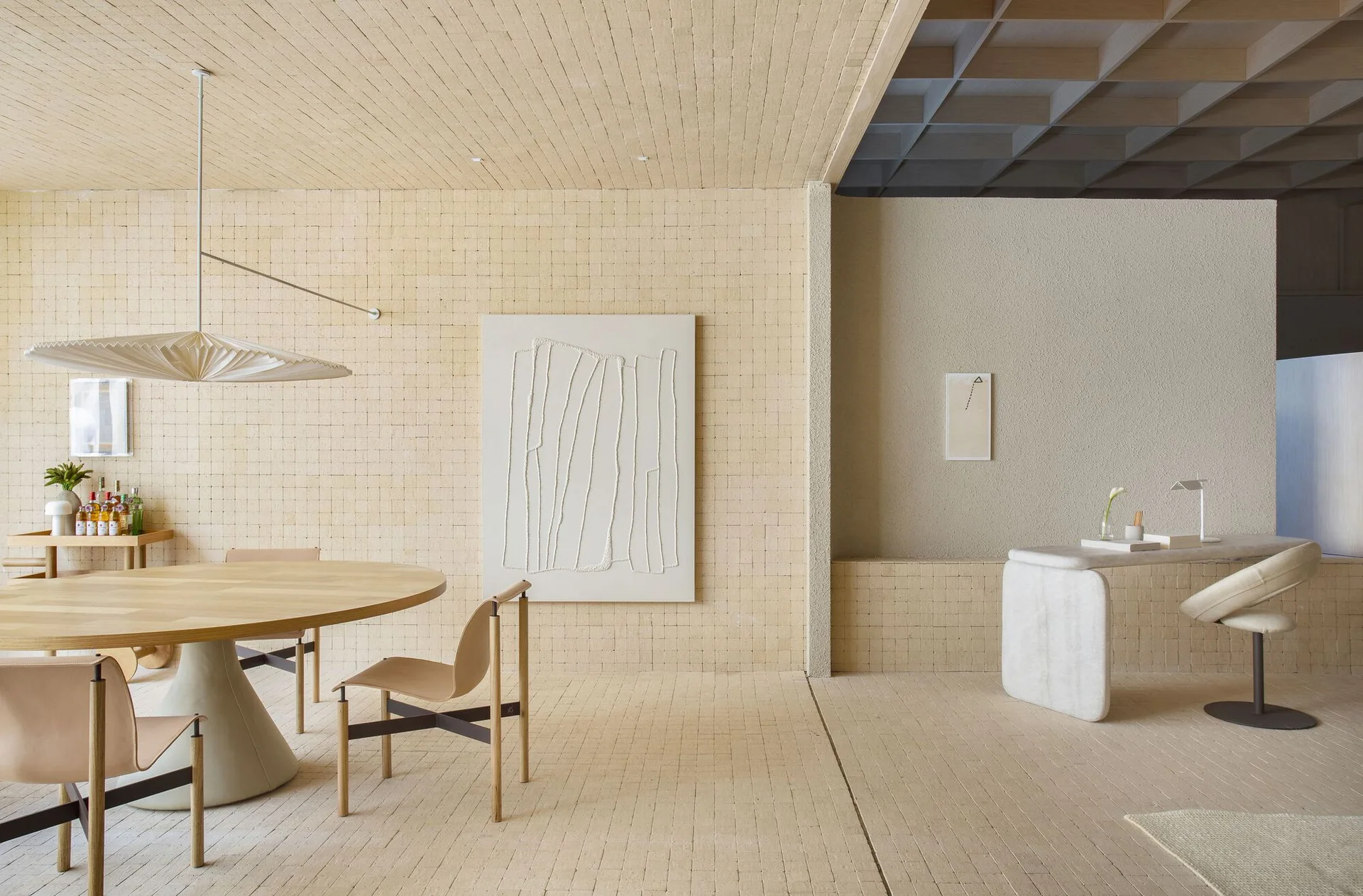A minimalist beige dining room with a round wooden table, leather sling chairs, textured tile walls, and a separate study area.