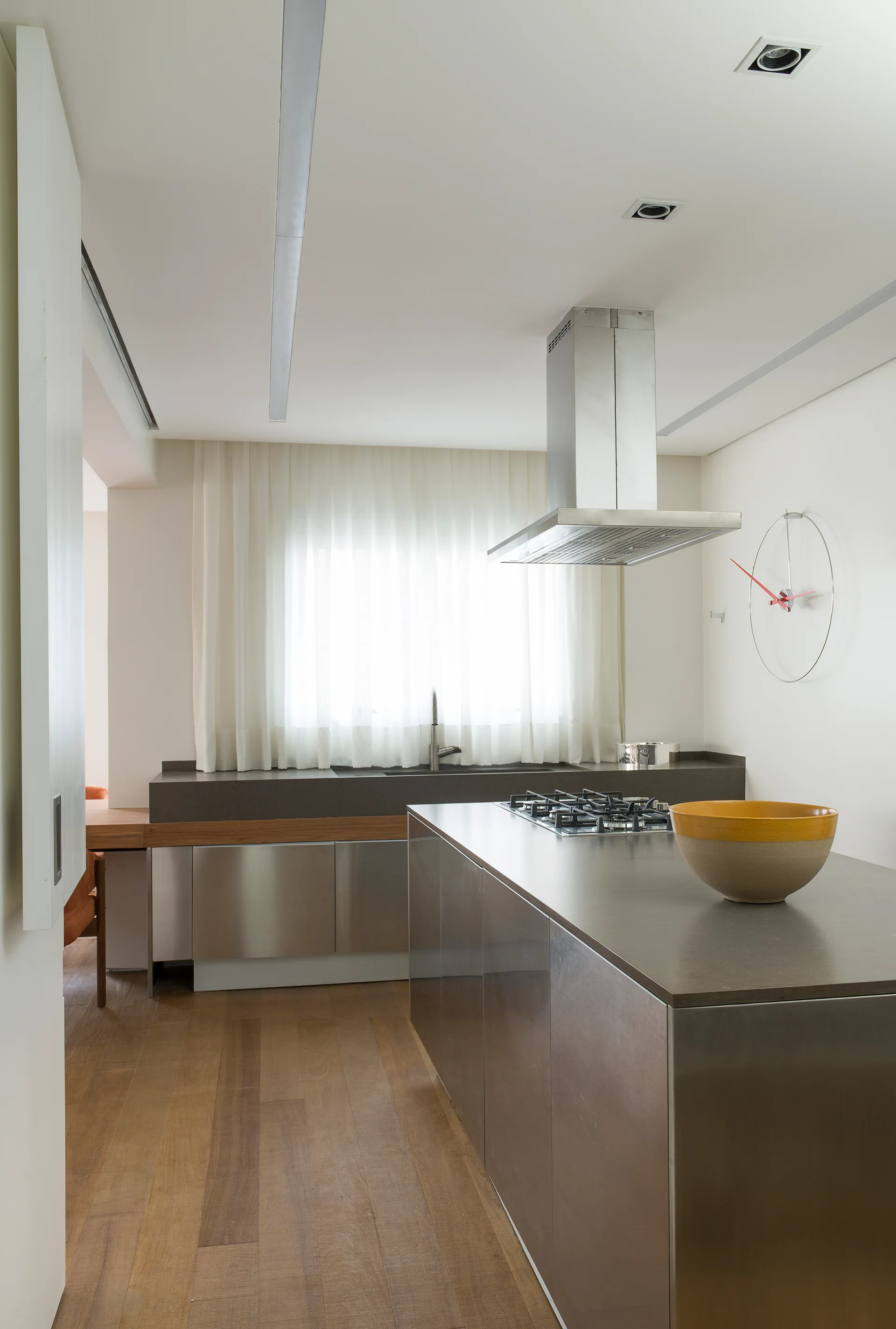 A minimalist kitchen features a central stainless steel island with a gas cooktop, matching silver cabinets, white walls, and warm wood plank flooring.