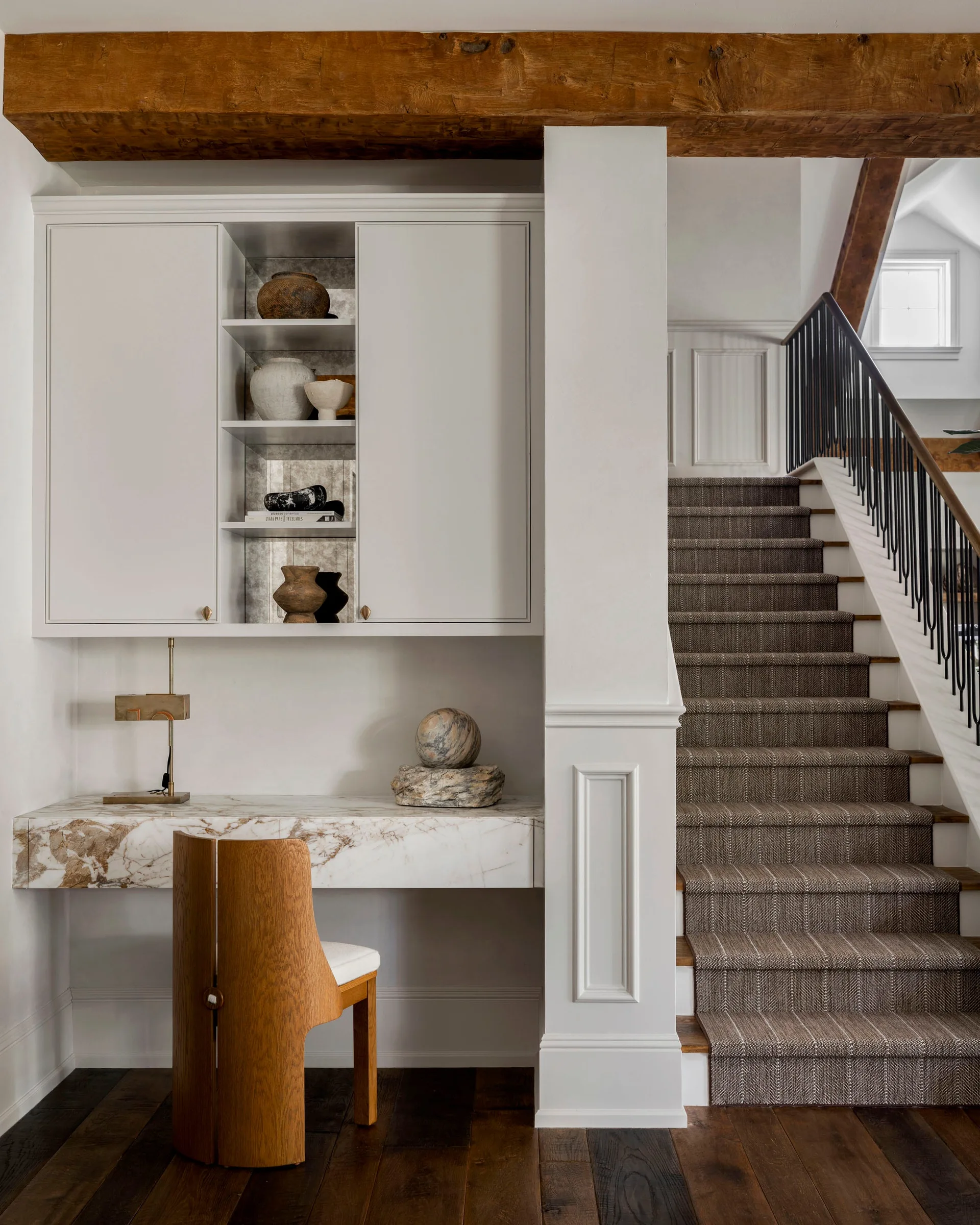 A built-in marble desk nook sits beside a staircase, featuring a curved wooden chair, cream-colored upper cabinets, and a textured stair runner.