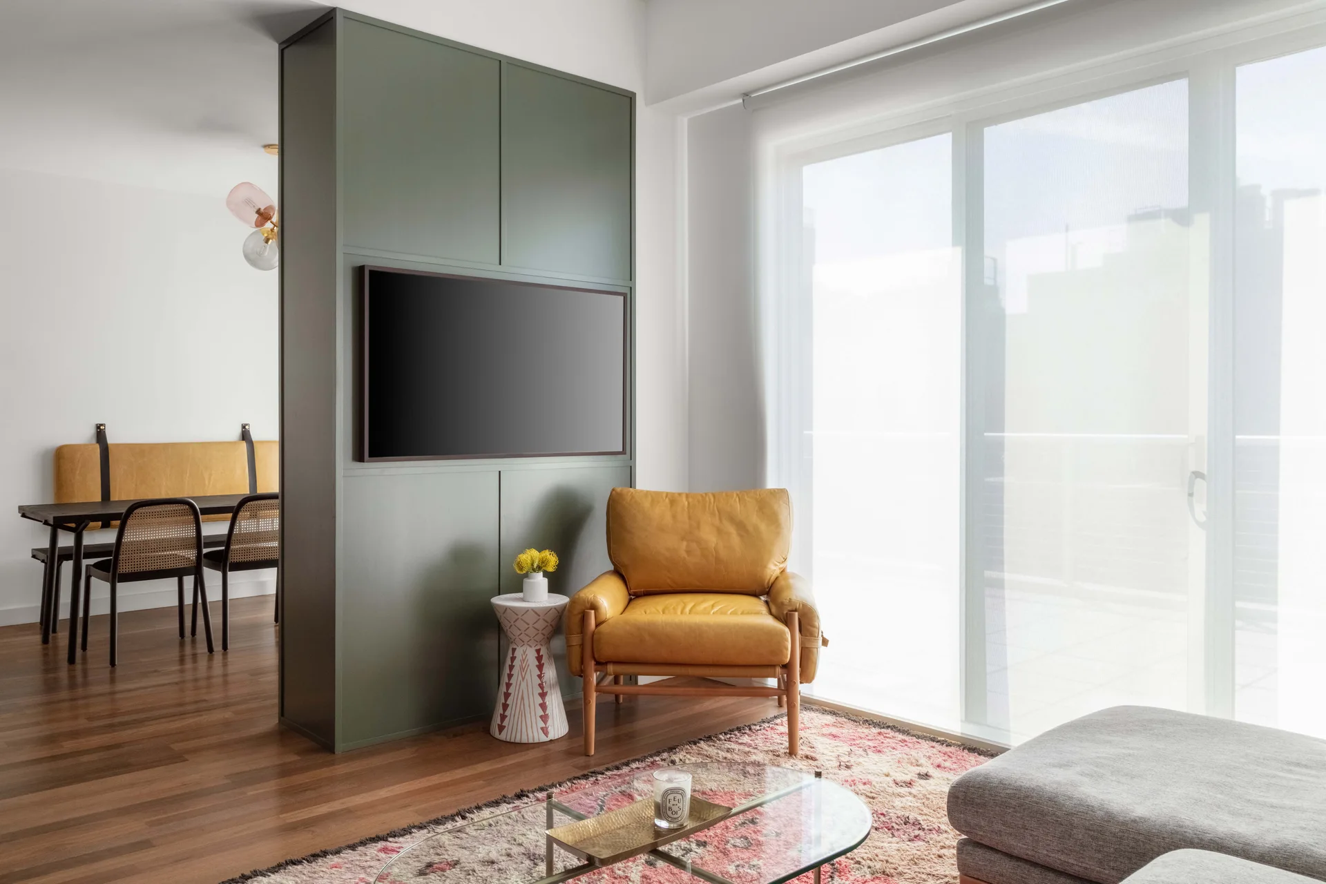 A modern living space features an olive green media partition wall, a mustard yellow leather armchair, a glass coffee table, and an adjacent dining area.