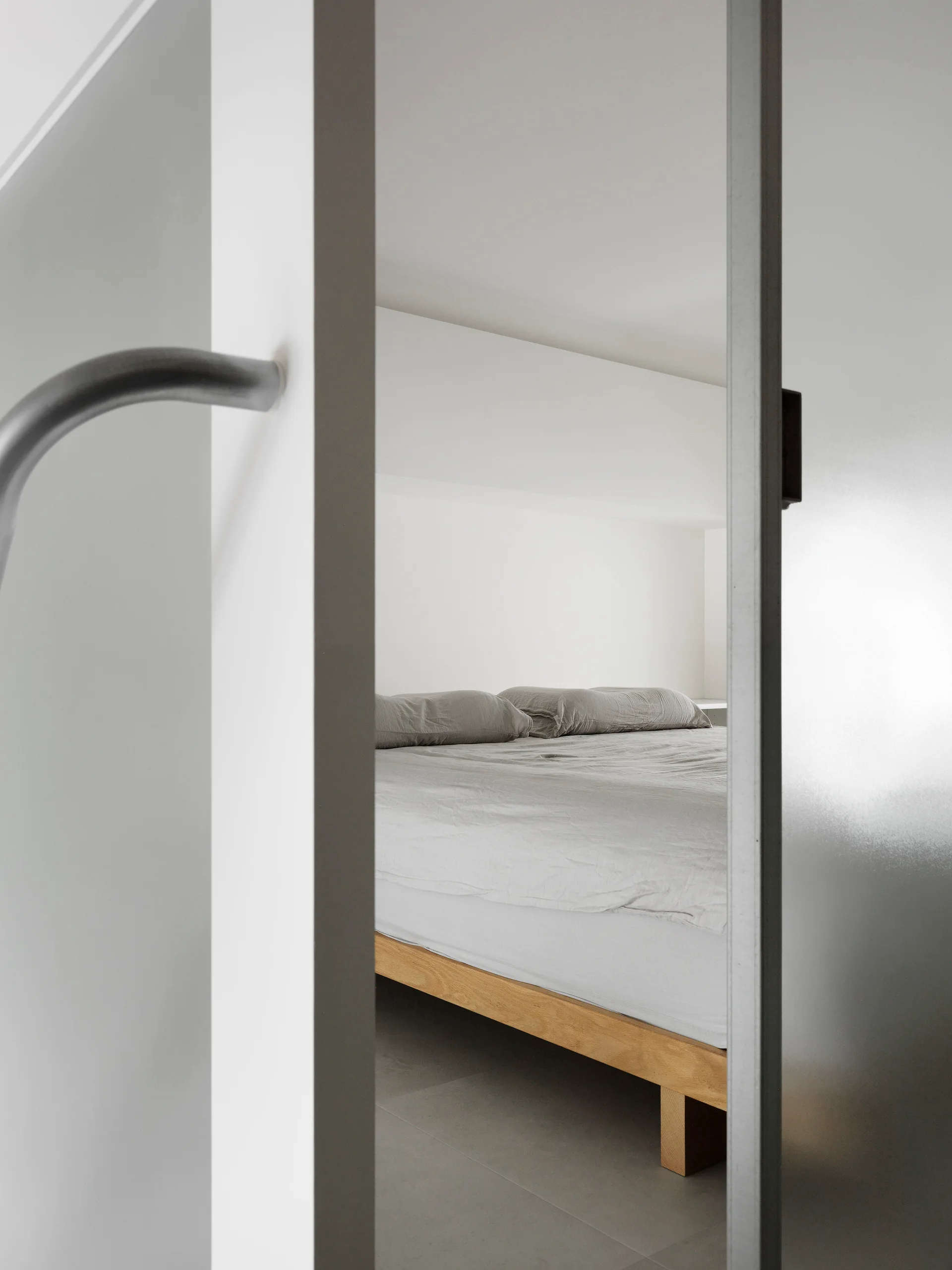 A view through a doorway shows a minimalist bedroom with a low wooden platform bed frame, grey linens, and simple white walls.
