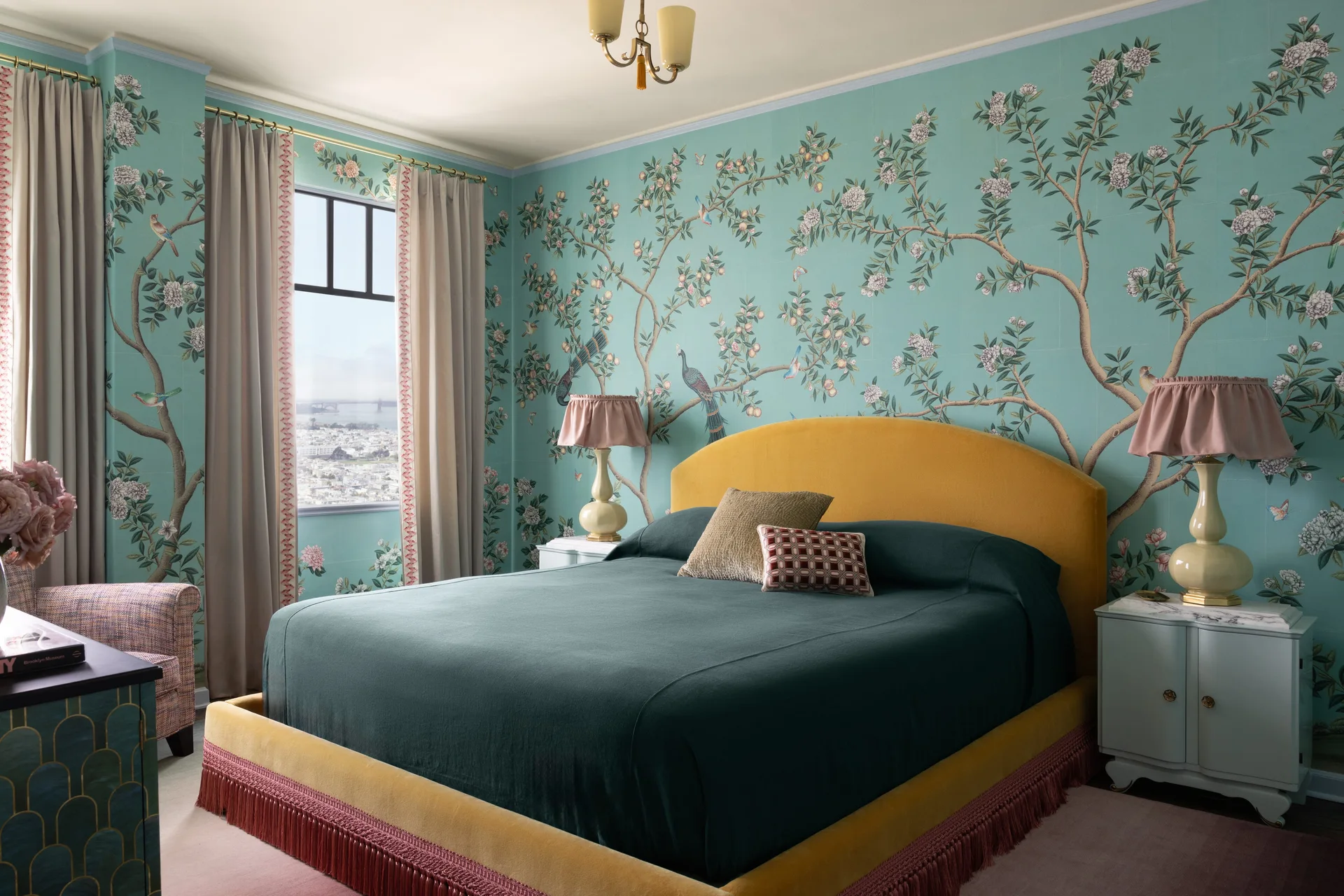 A bedroom features a mustard yellow upholstered bed with fringe trim set against teal floral wallpaper and flanked by light blue nightstands with lamps.
