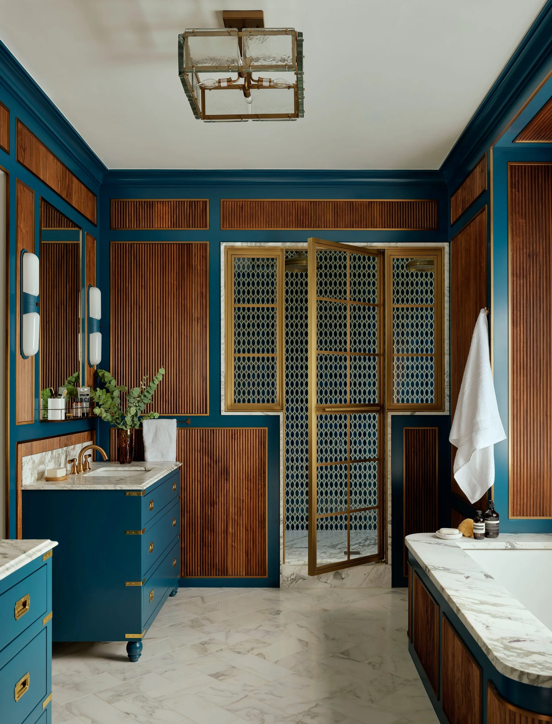 A blue bathroom features fluted wood-paneled walls, a marble-topped blue vanity, and a gold-framed glass shower door on a marble floor.