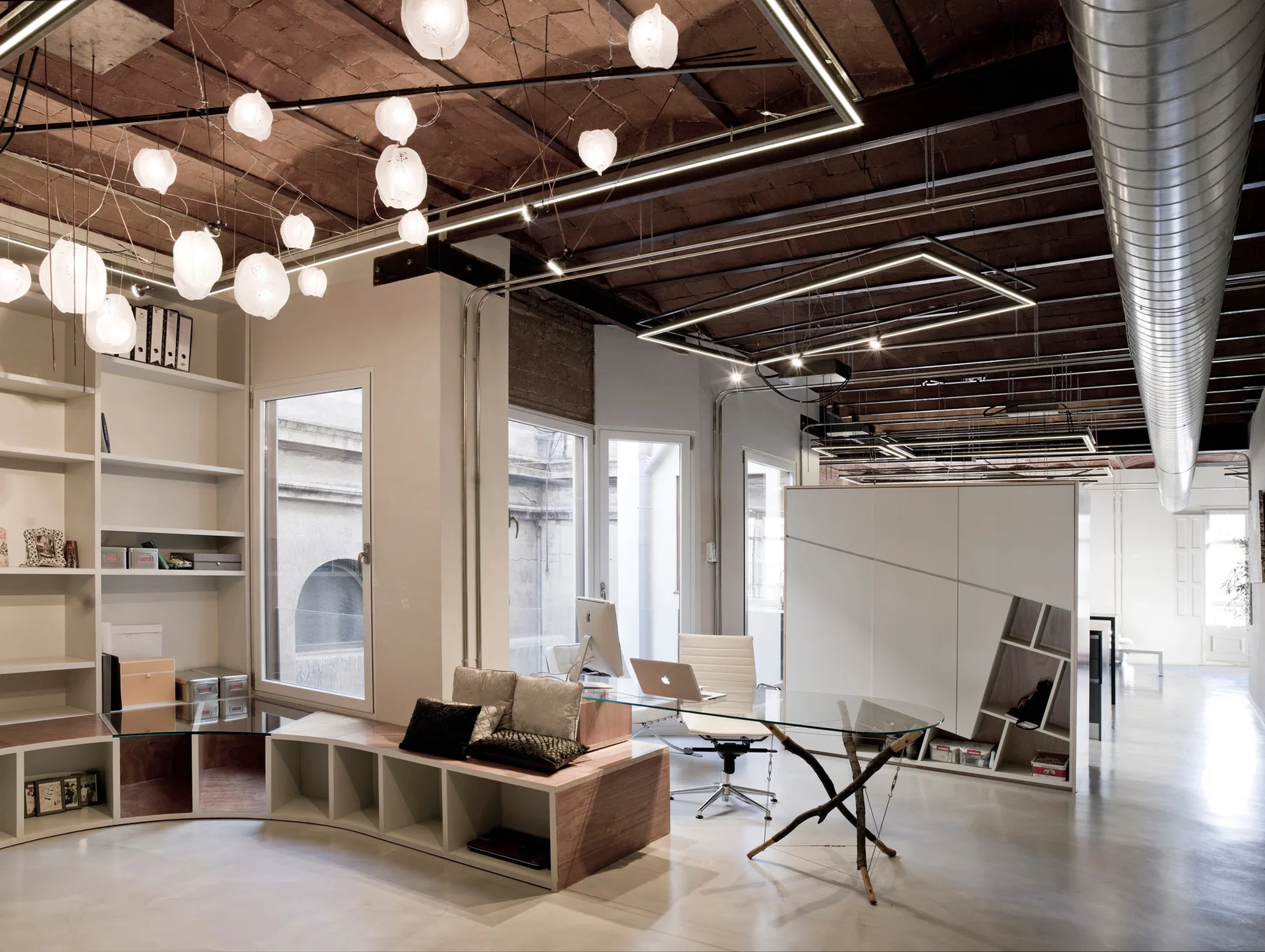 A modern office space featuring a glass-topped desk with a branch base, curved white shelving, and a vaulted brick ceiling.