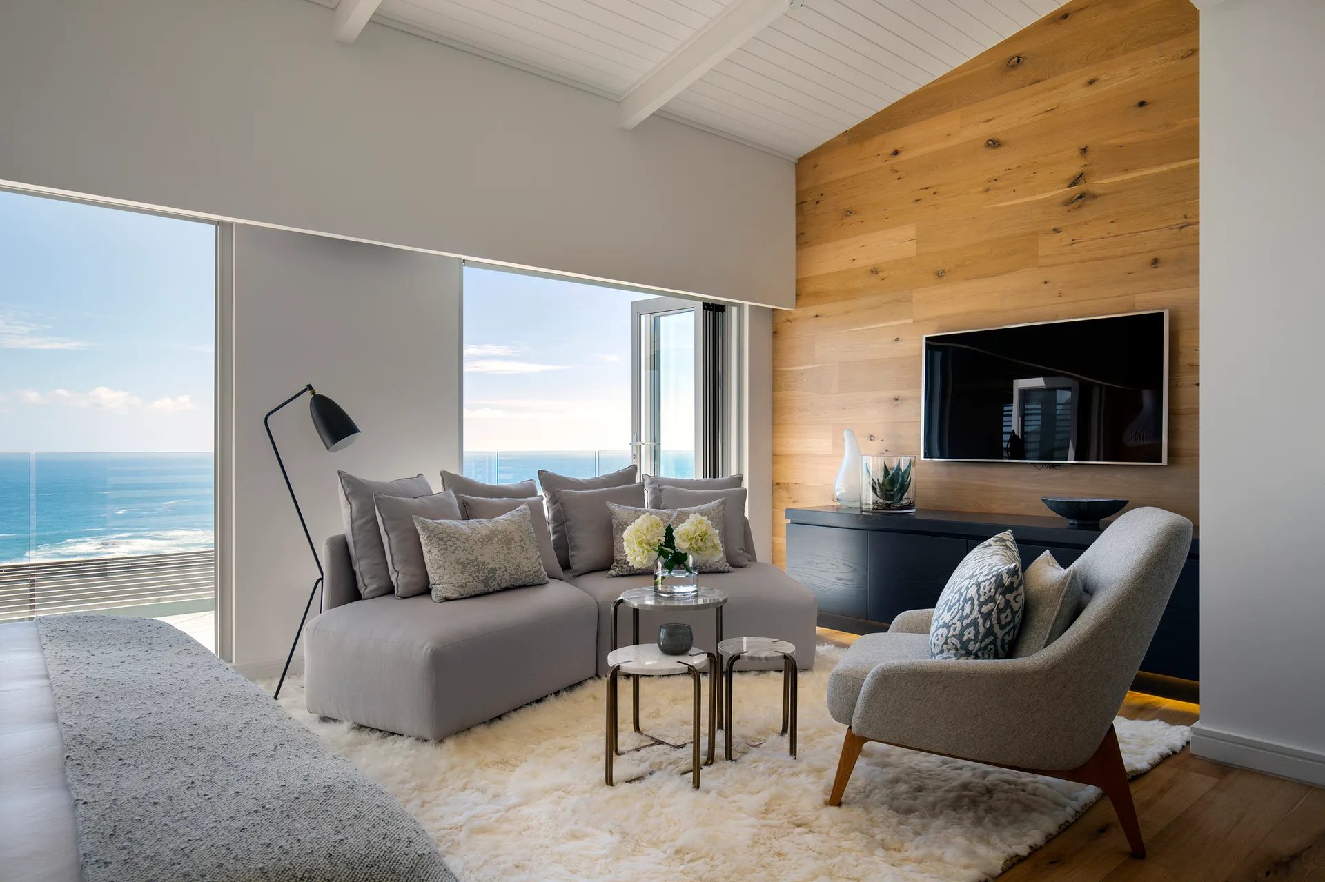 A modern living room features a grey sectional sofa, nesting tables, and a flat-screen TV mounted on a wood-paneled accent wall next to an ocean view.