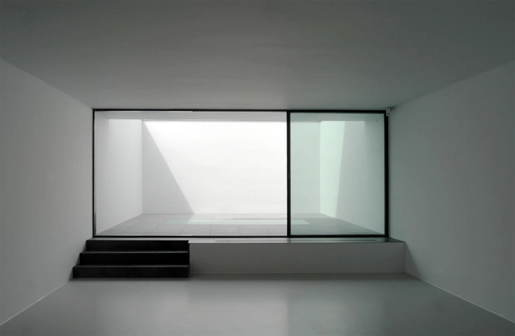A minimalist empty room features matte grey floors, white walls, and a large sliding glass door that opens to a bright courtyard with black concrete steps.