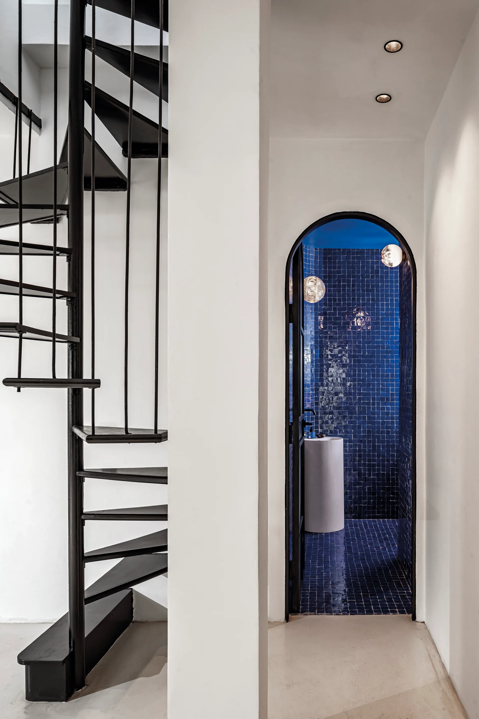 A black metal spiral staircase stands next to an arched doorway revealing a bathroom with glossy dark blue tiles and a white pedestal sink.