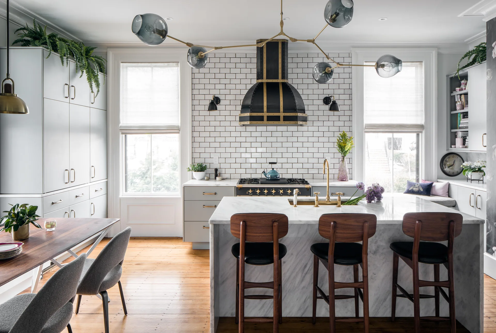 A modern kitchen featuring a marble waterfall island with wood stools, a black and brass range hood, and white subway tile backsplash.