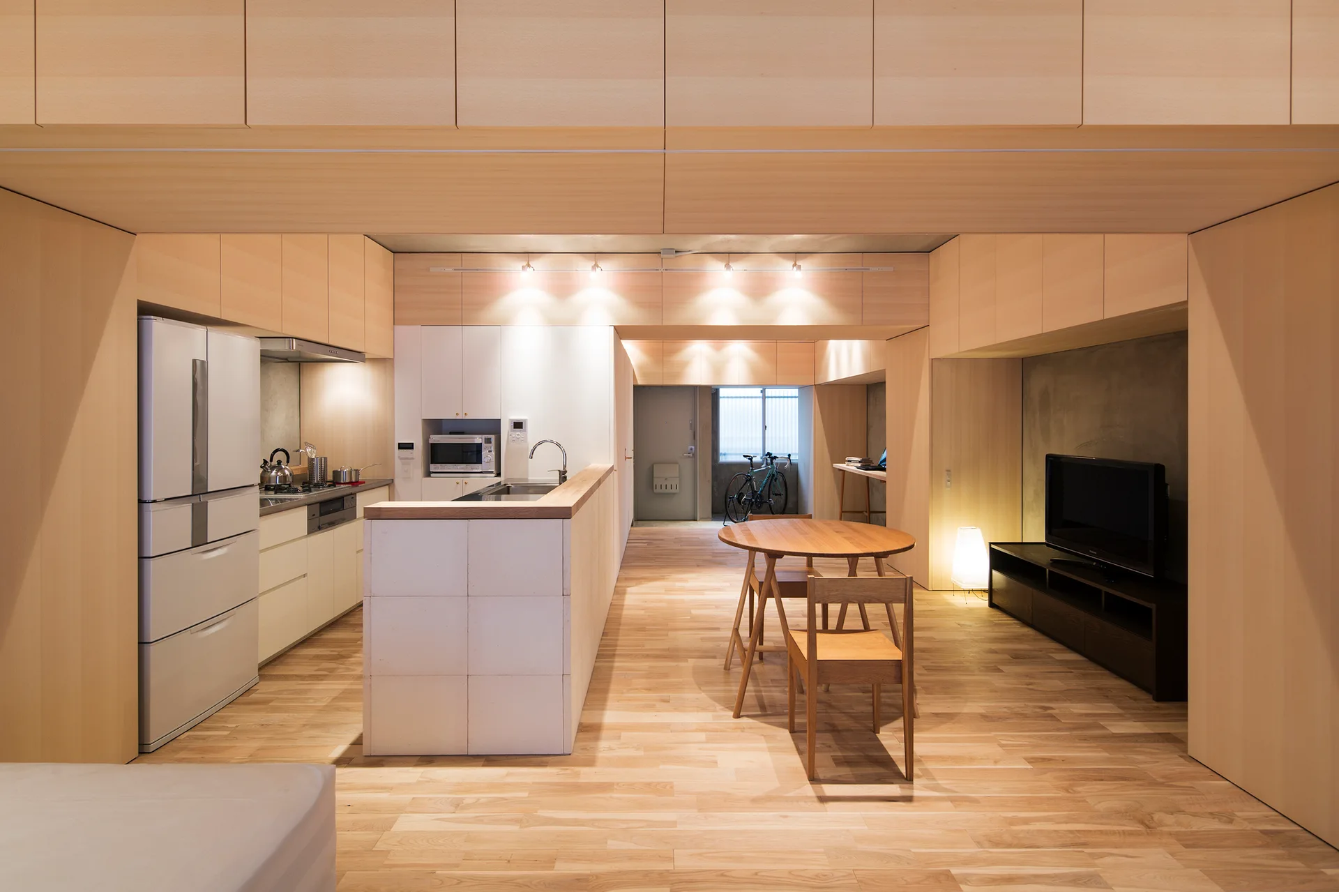 A minimalist apartment interior features light wood wall cladding, a compact dining set, and a white tile kitchen island next to a modern refrigerator.