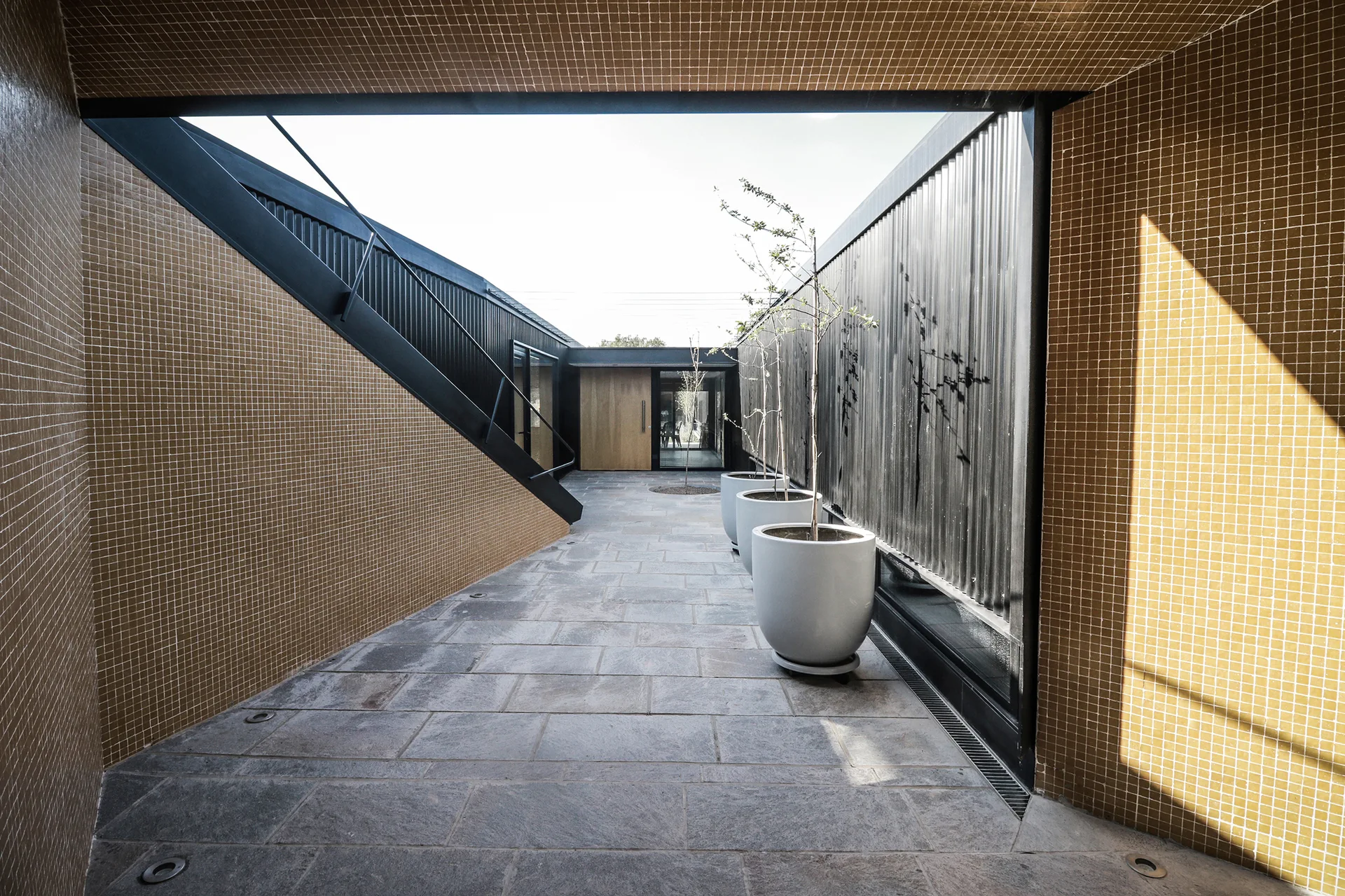 An outdoor courtyard features yellow-brown tiled walls, a black corrugated metal wall, grey stone pavers, and three large grey planters.
