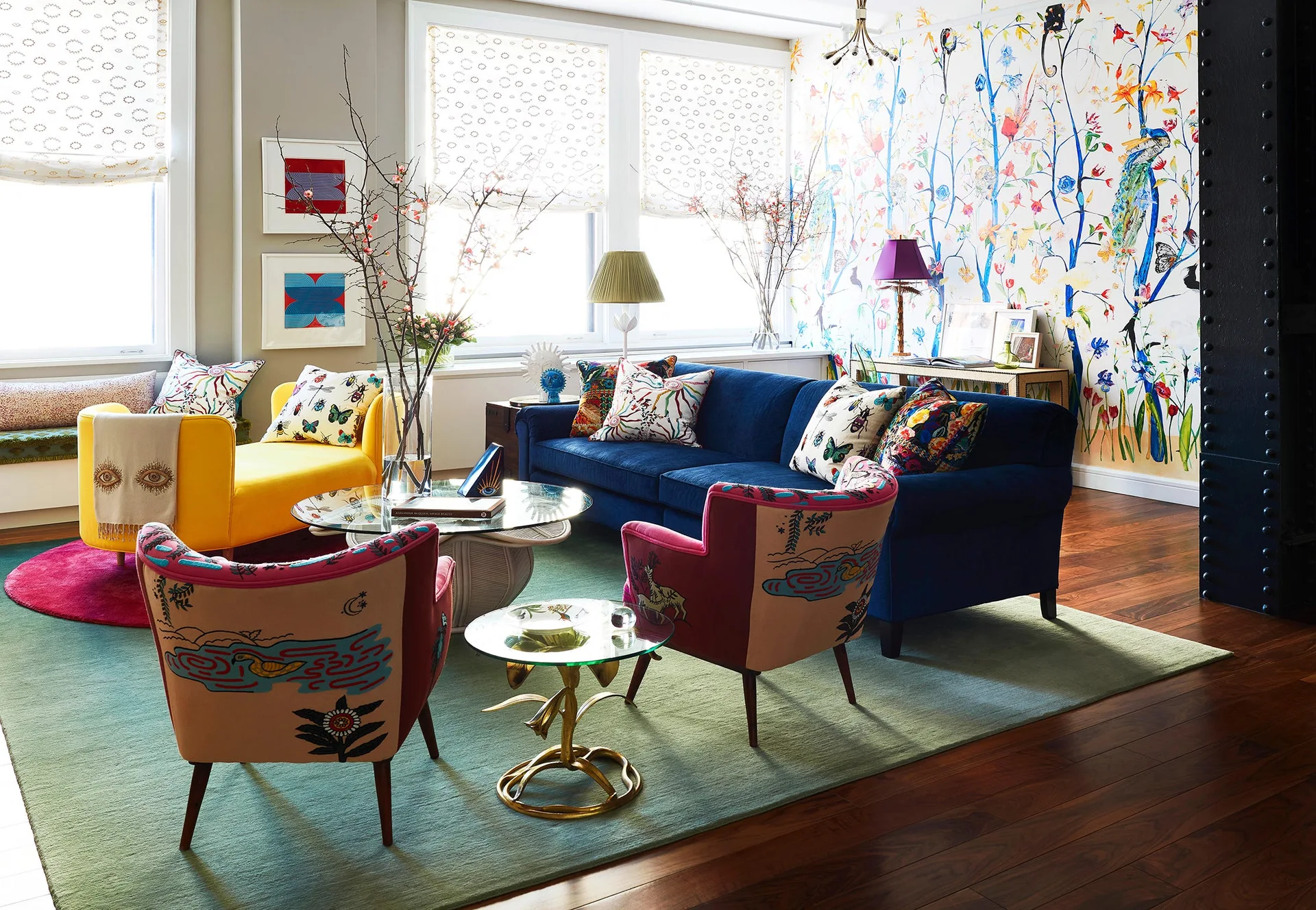 An eclectic living room with a blue sofa, a yellow chaise, floral wallpaper, patterned armchairs, and a green area rug on wood floors.
