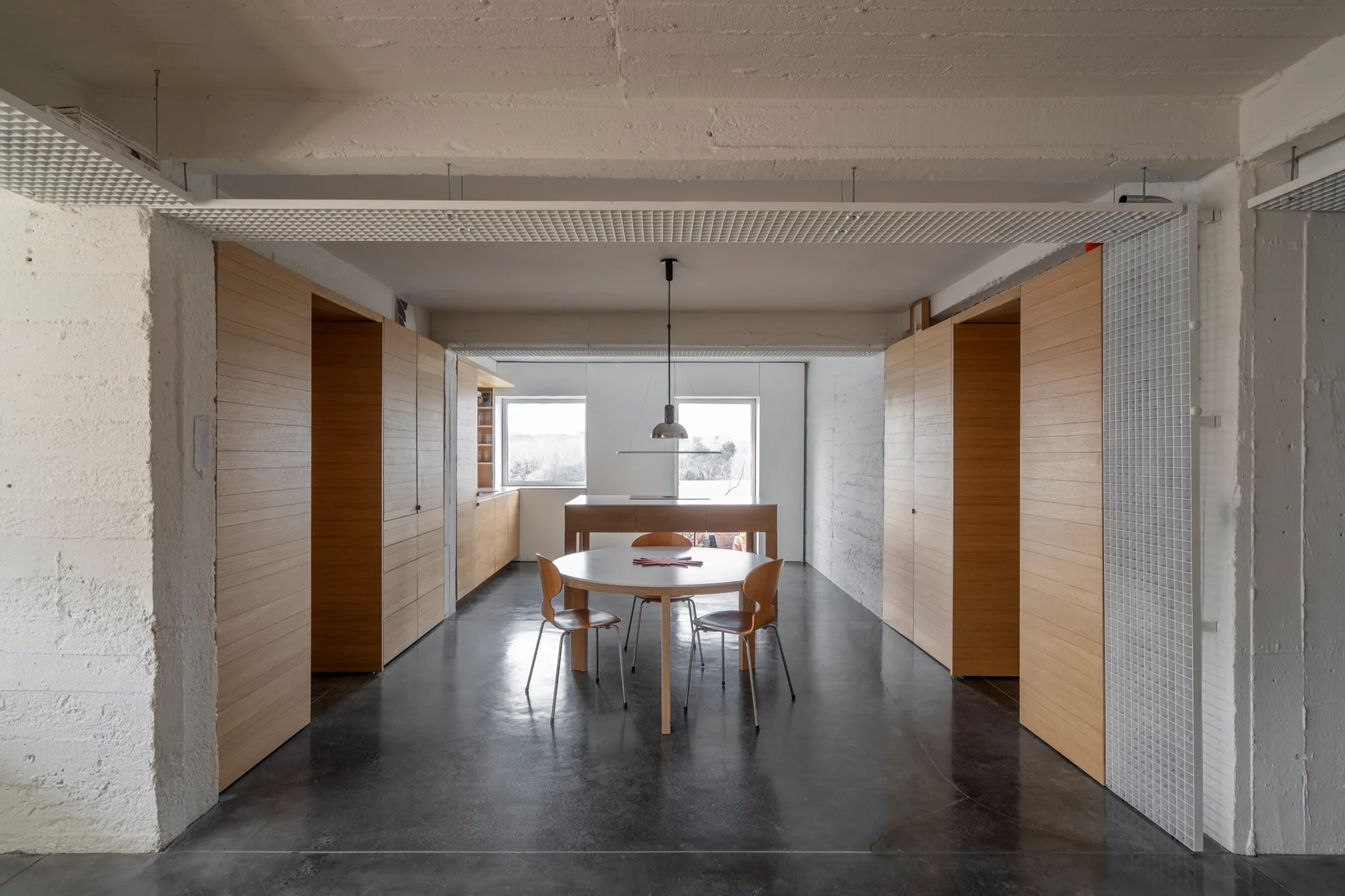 A minimalist dining room features a round table with wooden chairs, flanked by large wood-paneled storage units on a dark concrete floor.