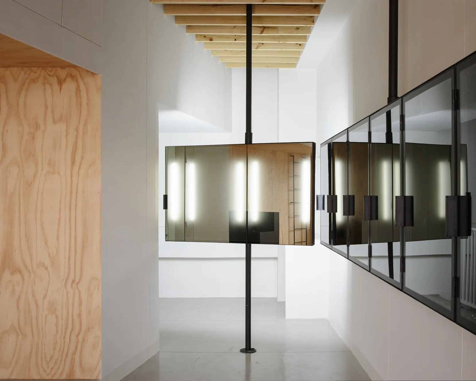 A minimalist hallway features mirrors mounted on vertical black poles, integrated light strips, white walls, and exposed wooden ceiling beams.