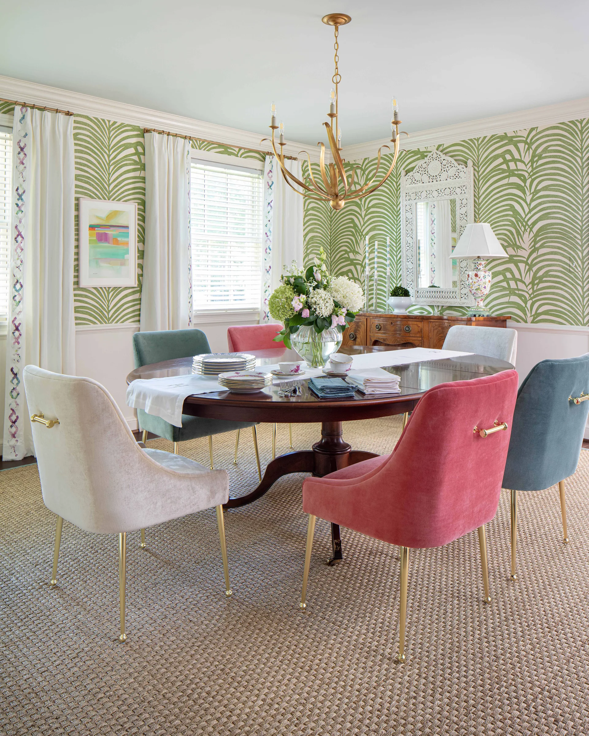 A round wooden dining table surrounded by colorful velvet chairs in a dining room with green palm-leaf wallpaper and a woven rug.